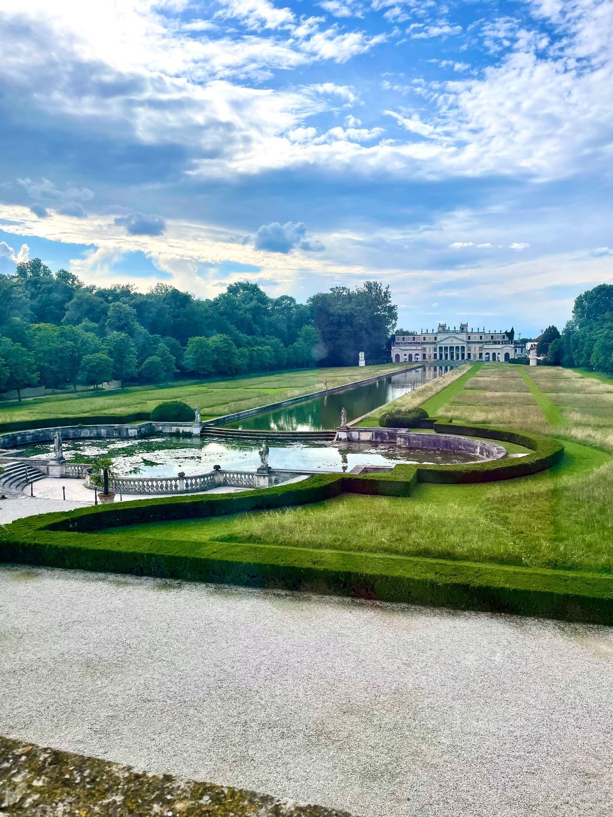Formal garden with reflecting pools, manicured lawns, trees, and a distant stately building under a cloudy sky