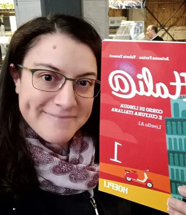 Woman with glasses holding a red “Feria” sign, indoors, smiling at the camera.