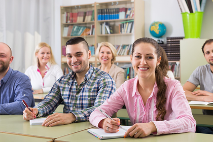 People seated at desks in a bright classroom, smiling and writing in notebooks