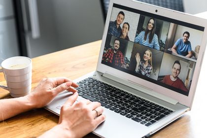 Video call on a laptop with six participants, hands typing at a desk beside a coffee cup.