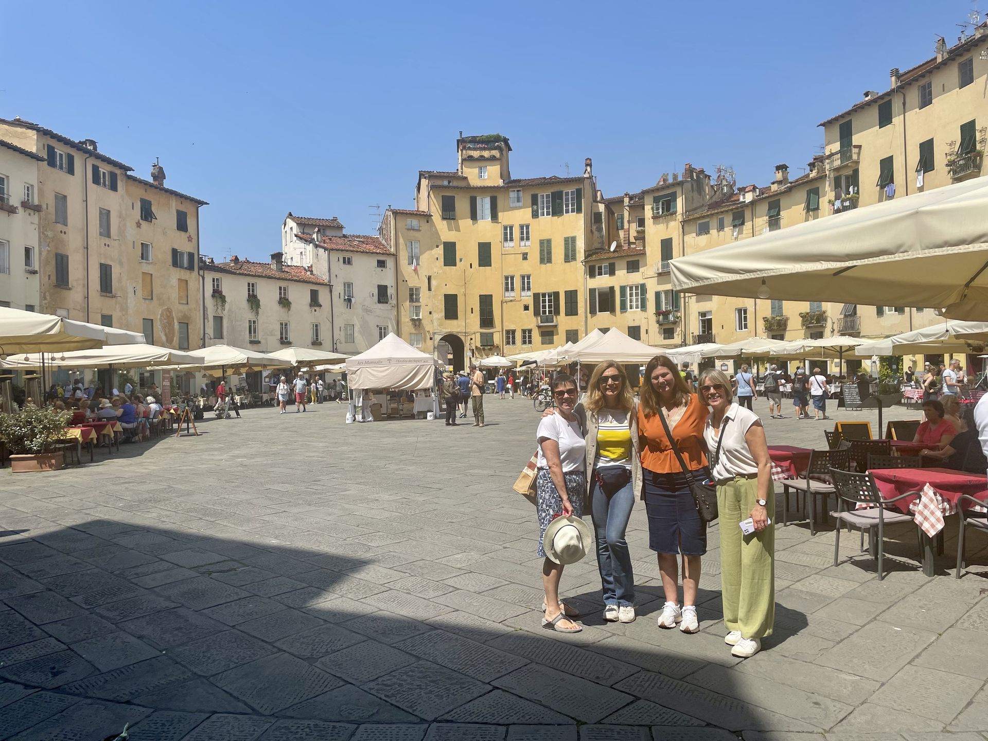 Tourists walking in a sunny European plaza lined with cafes and historic yellow buildings