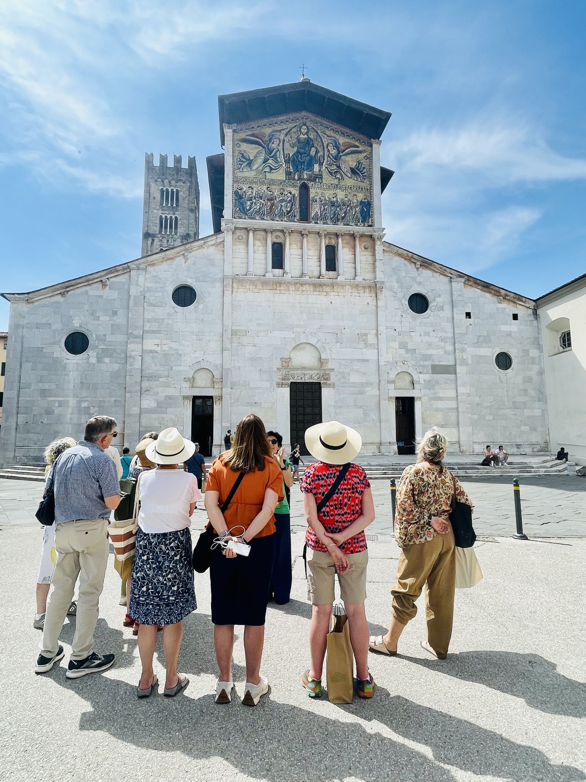 Tourists in hats stand in a plaza facing an ornate white stone church with a tower and blue sky.