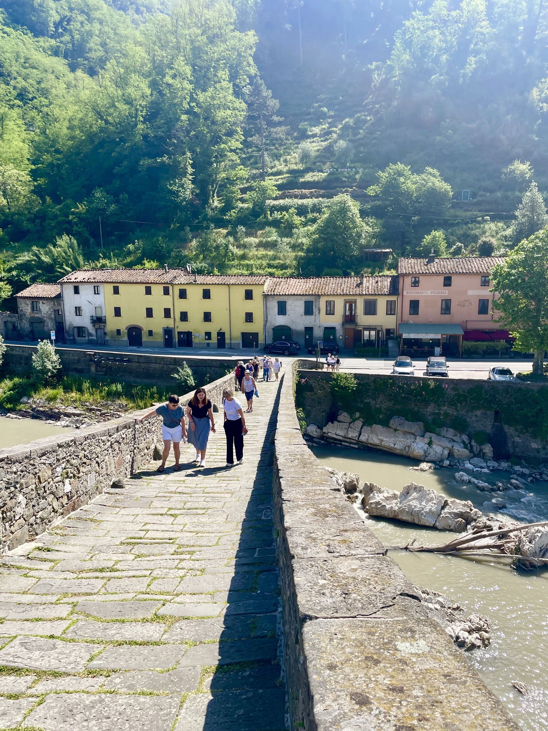 Stone bridge to colorful riverside houses with people walking, surrounded by green hillside.
