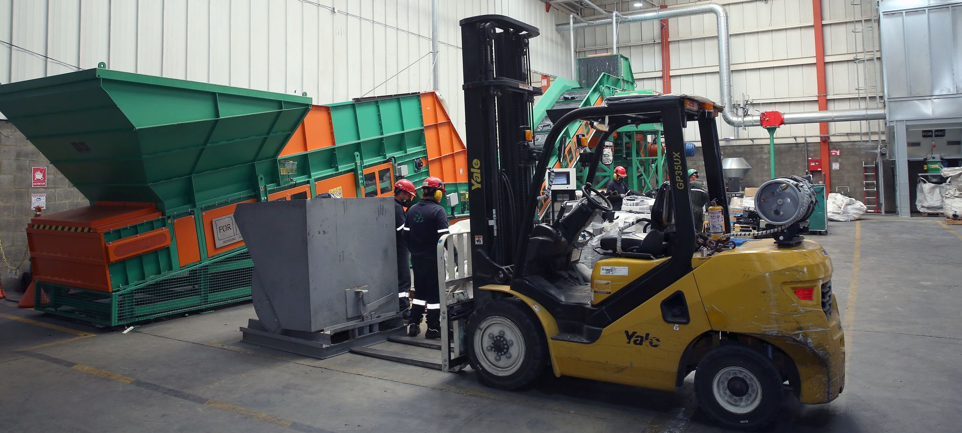 Una carretilla elevadora amarilla descarga una tolva industrial verde-anaranjada en un almacén.