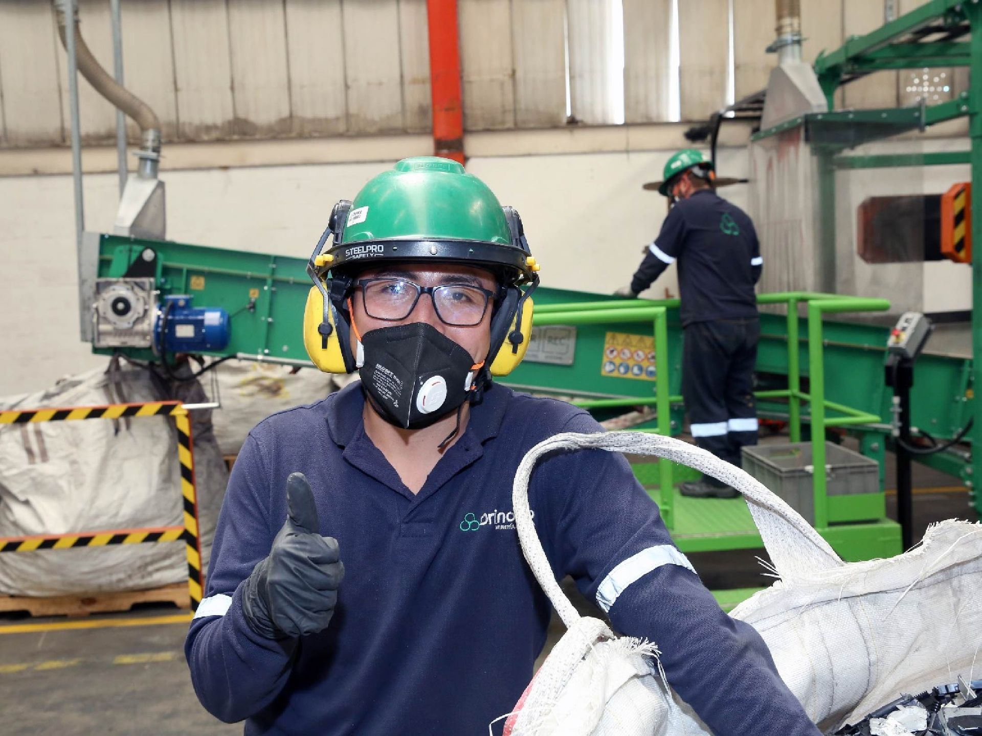 Un trabajador con casco verde y mascarilla levanta el pulgar en una fábrica junto a maquinaria industrial.