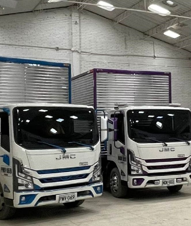 Dos camiones de reparto JMC blancos estacionados dentro de un almacén, uno con detalles azules y otro con detalles morados.