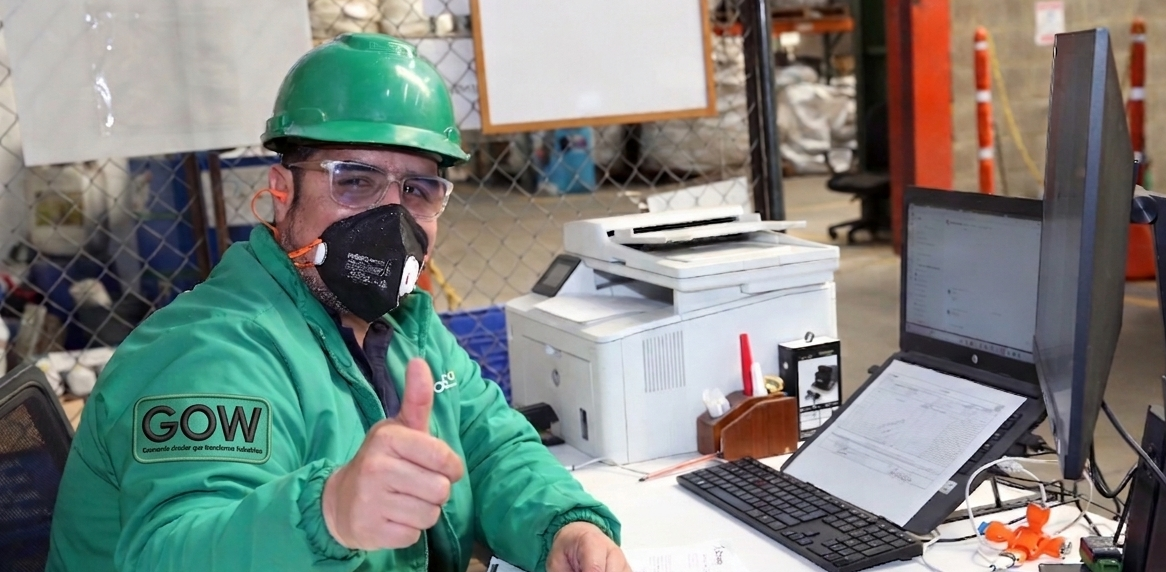 Un trabajador con equipo de seguridad verde levanta el pulgar junto a una computadora portátil en un espacio de trabajo industrial.