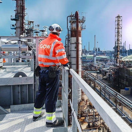 Un trabajador con chaleco de seguridad naranja observa una refinería industrial desde una plataforma en la azotea.
