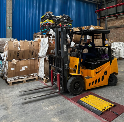 Una carretilla elevadora amarilla transporta cajas apiladas en un almacén junto a palés y una rampa de acceso al suelo.