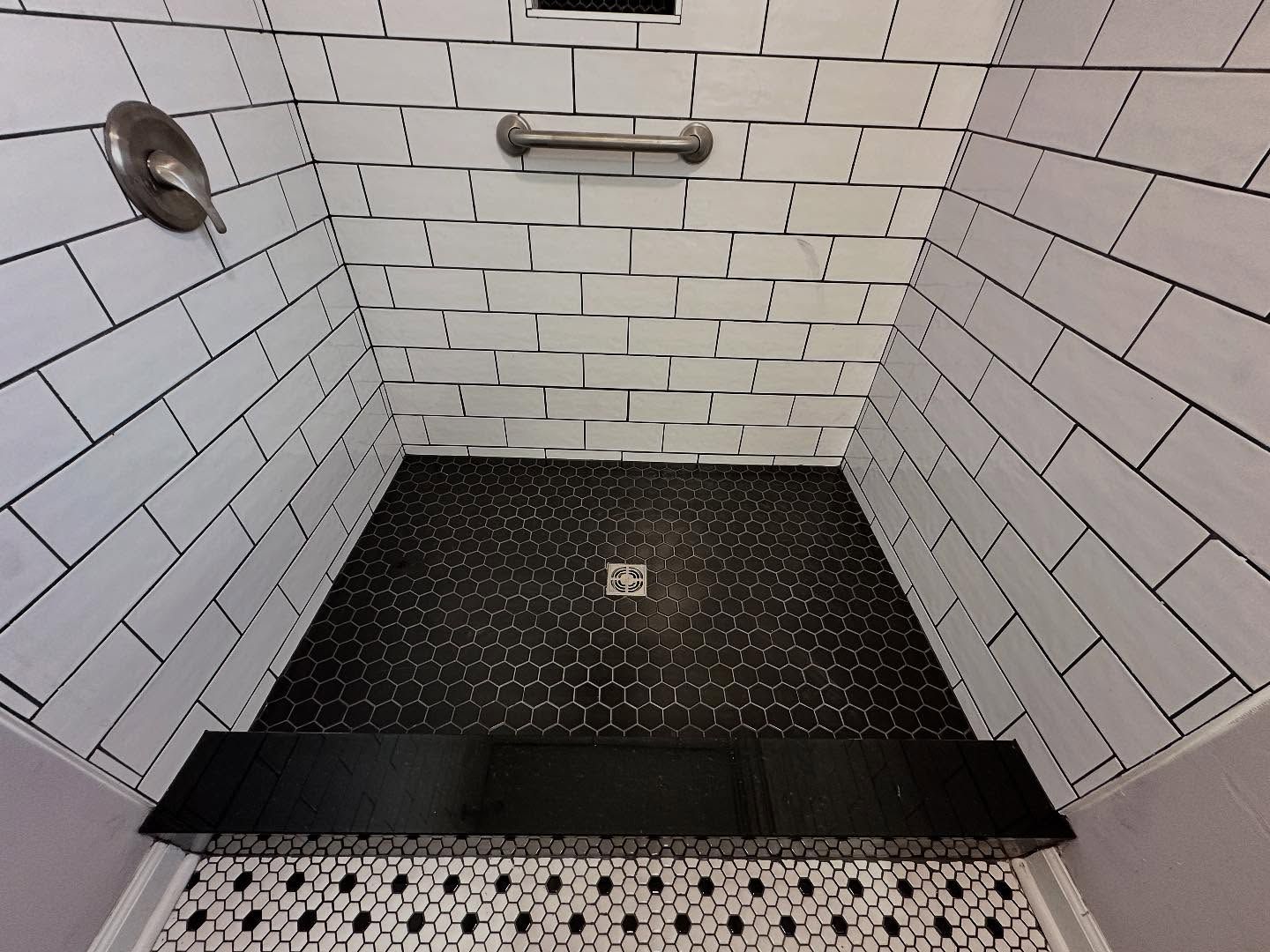 Empty tiled shower stall with black floor mat, drain, and a grab bar on the wall