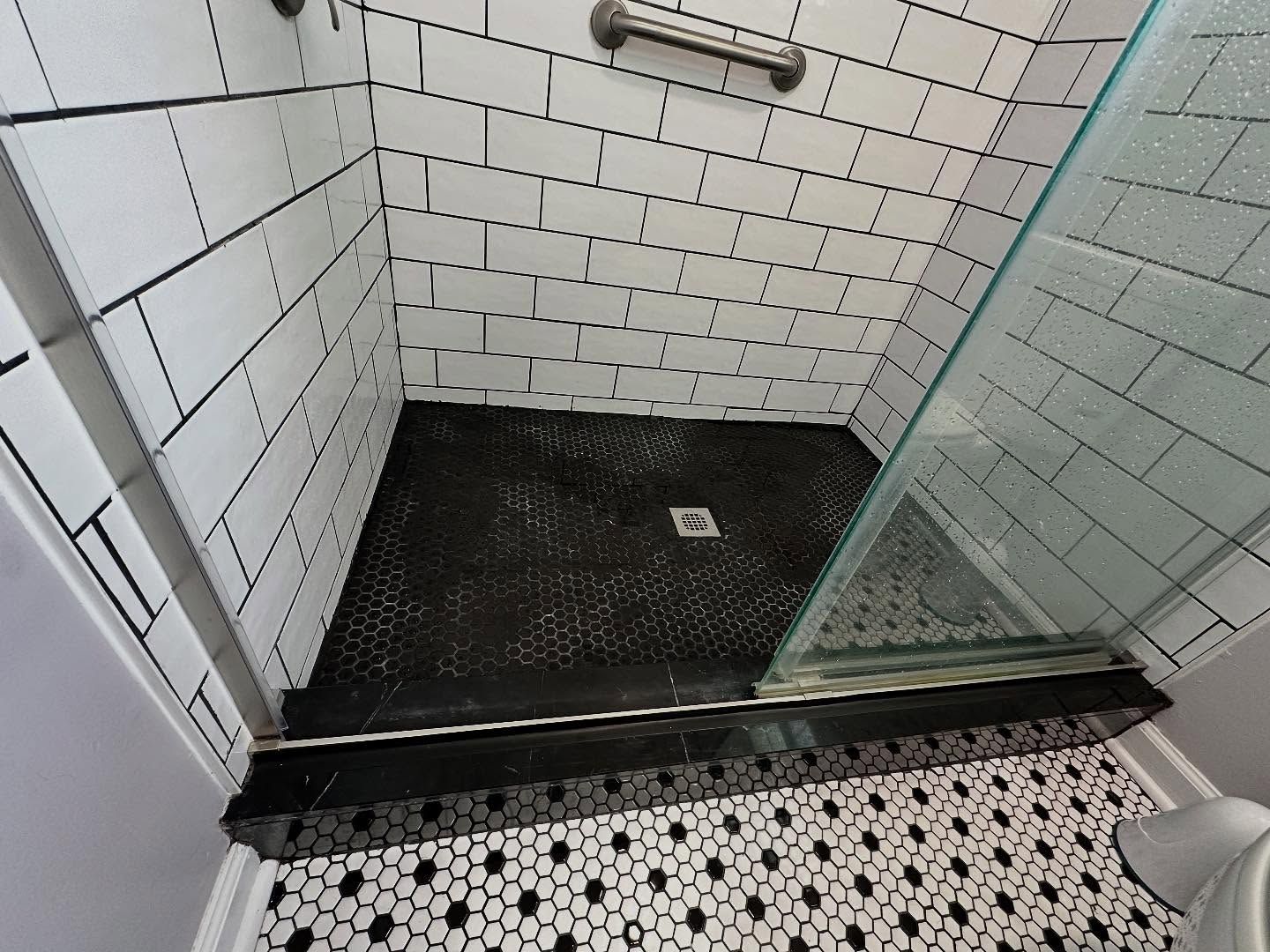 Small tiled shower stall with black floor pan and glass door, viewed from the entrance.