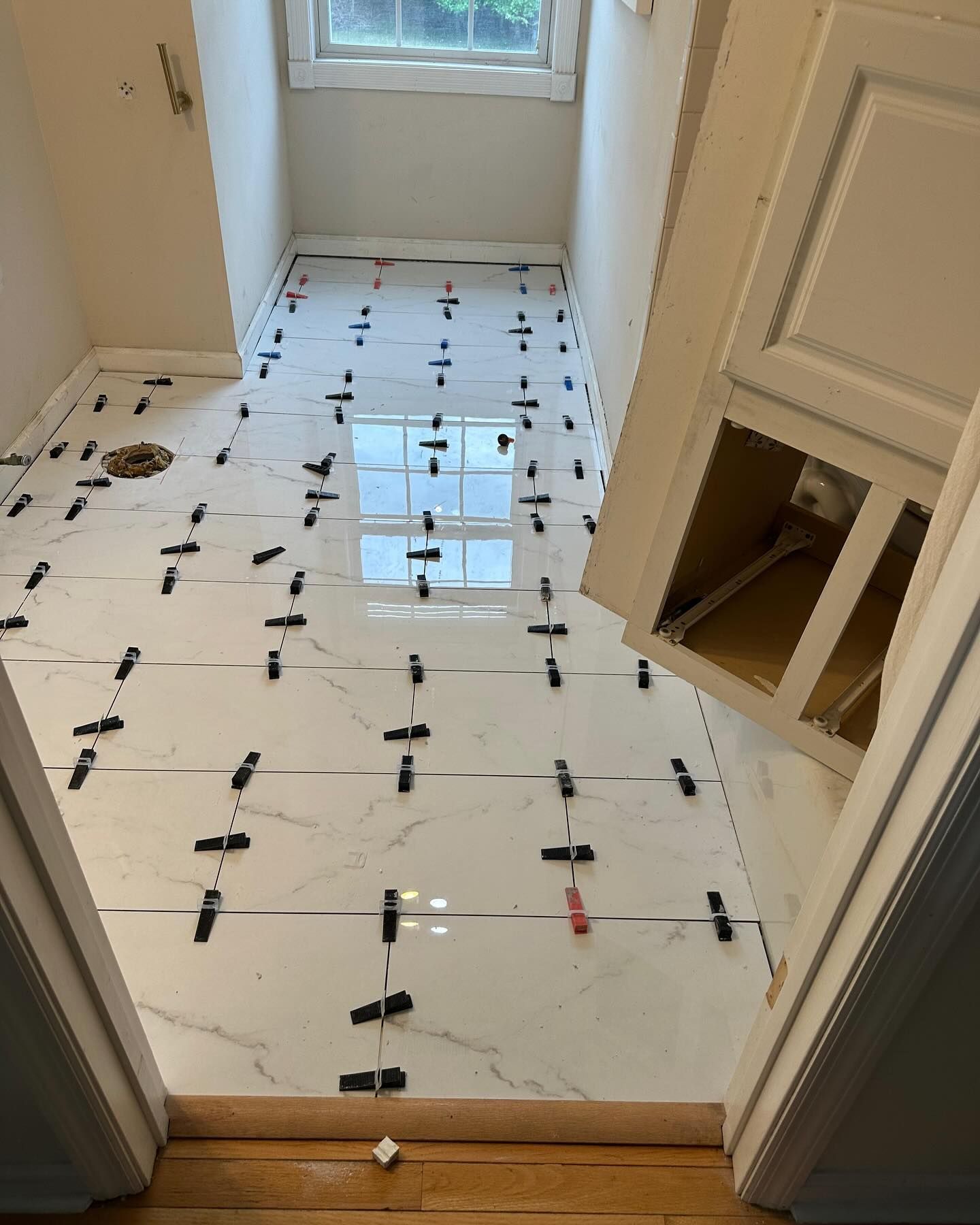 Tiled bathroom floor under renovation, with black leveling clips and a window ahead.