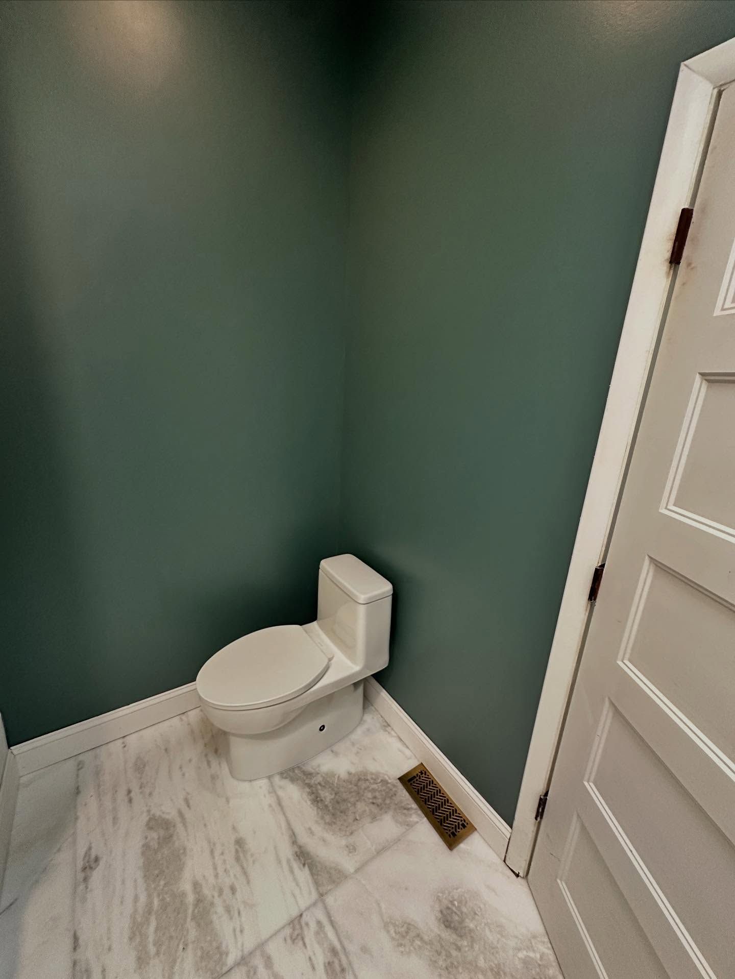 Small bathroom with a white toilet, green walls, marble-look floor, and a floor vent near the door.