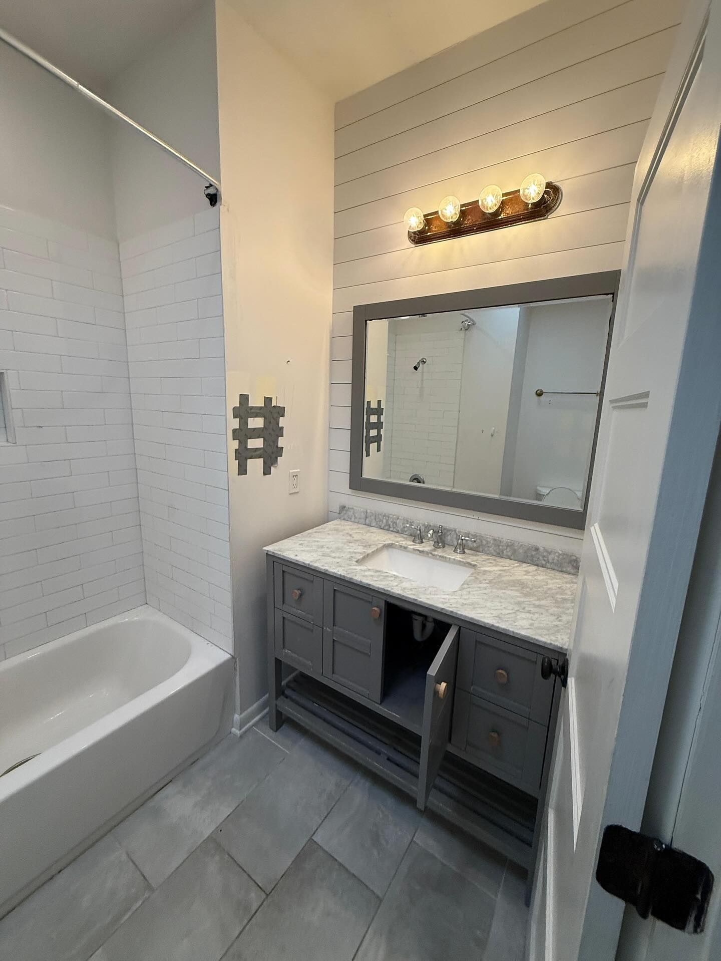 Small bathroom with white tub, gray vanity, large mirror, and wall light over beige tile.