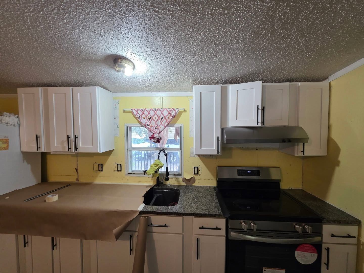 Small kitchen with white cabinets, black stove and countertop, yellow walls, and a window over the sink.