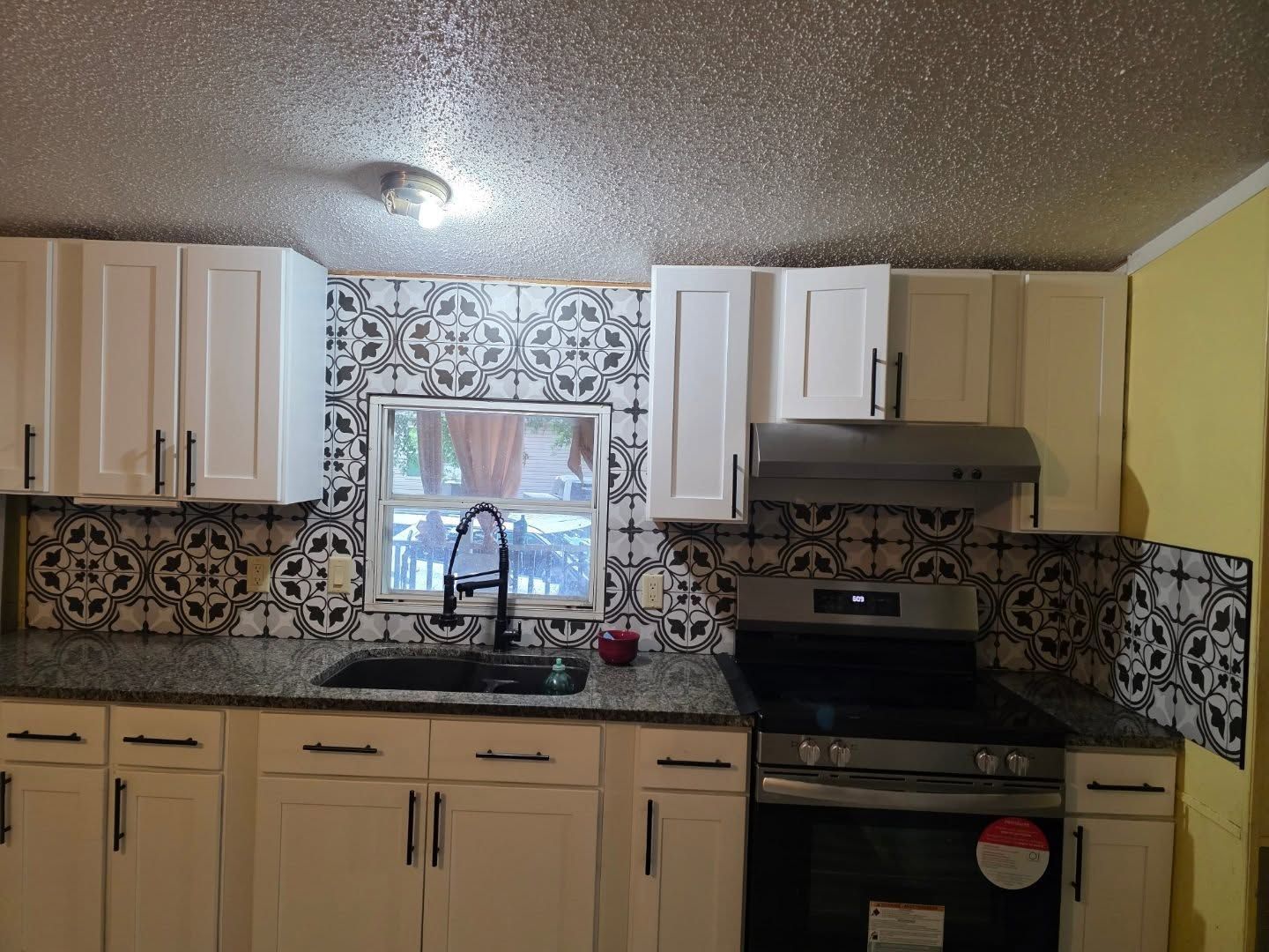 Small kitchen with beige cabinets, patterned backsplash, stainless sink, and black stove under bright ceiling light.
