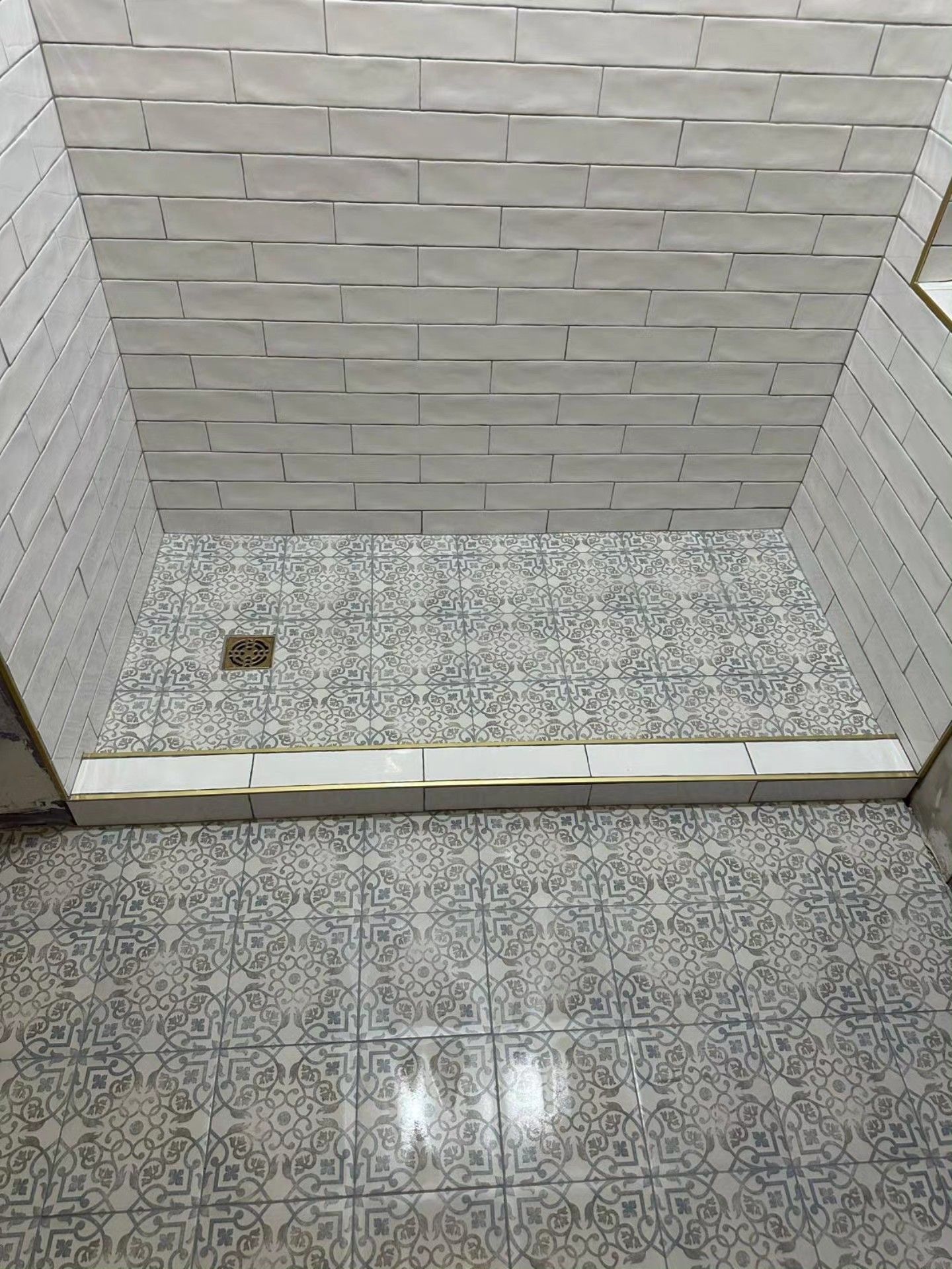 Small tiled restroom floor with a low step, drain, and white-and-gray wall tiles.