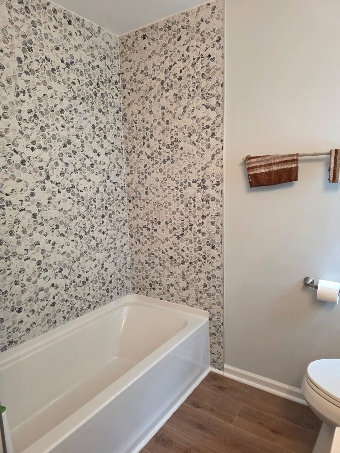 Bathtub in a tiled bathroom with patterned wall and wood-look floor, next to a toilet and towel rack