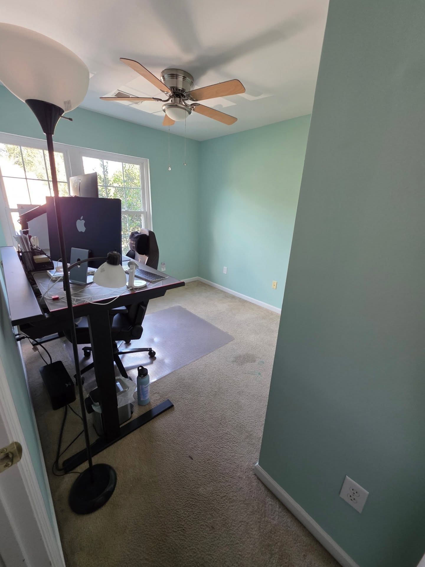 Home office with desk, computer monitors, carpet, teal walls, and a ceiling fan near a window.