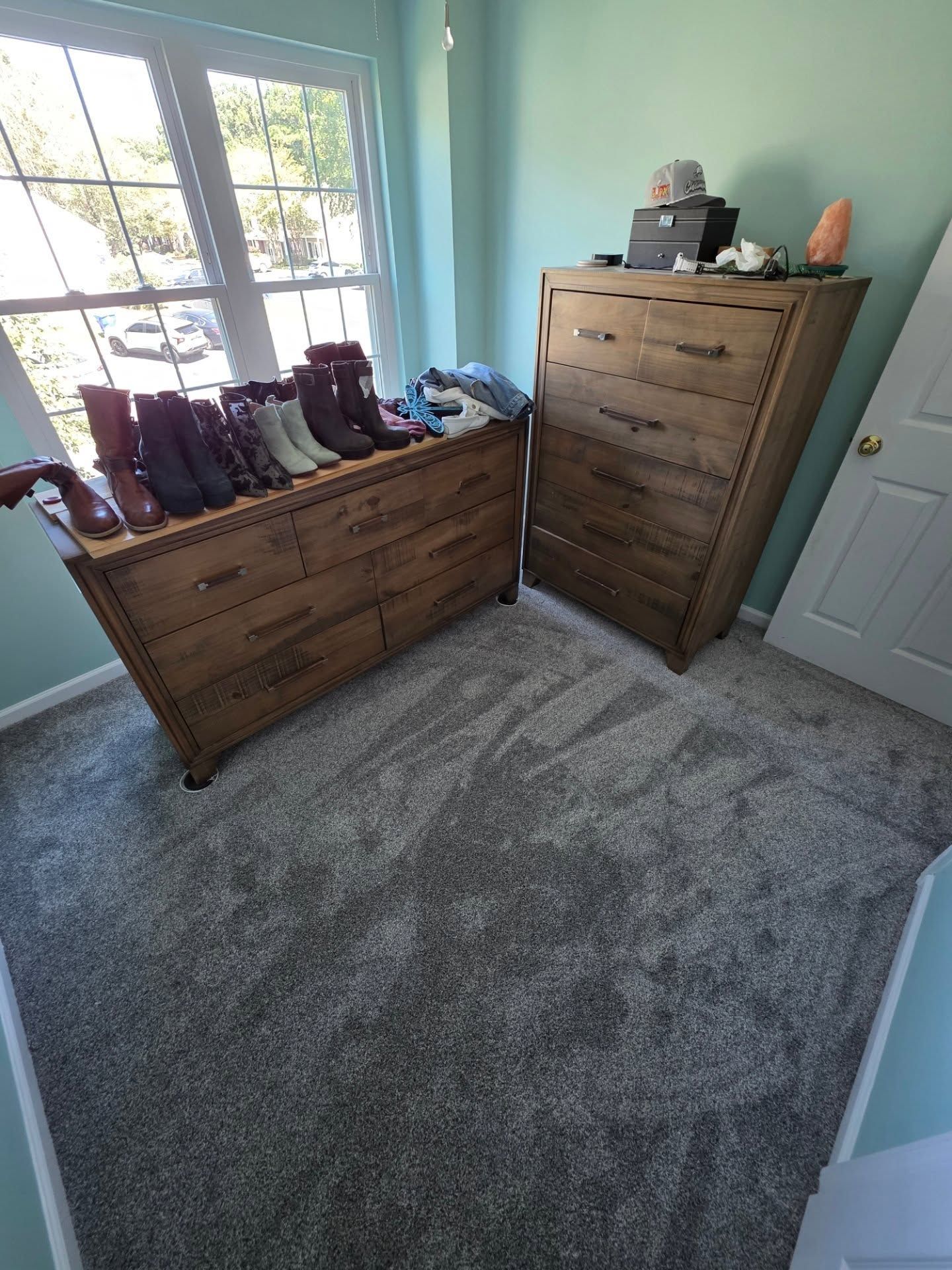 Bedroom corner with two wooden dressers, boots on one, gray carpet, and a window letting in daylight