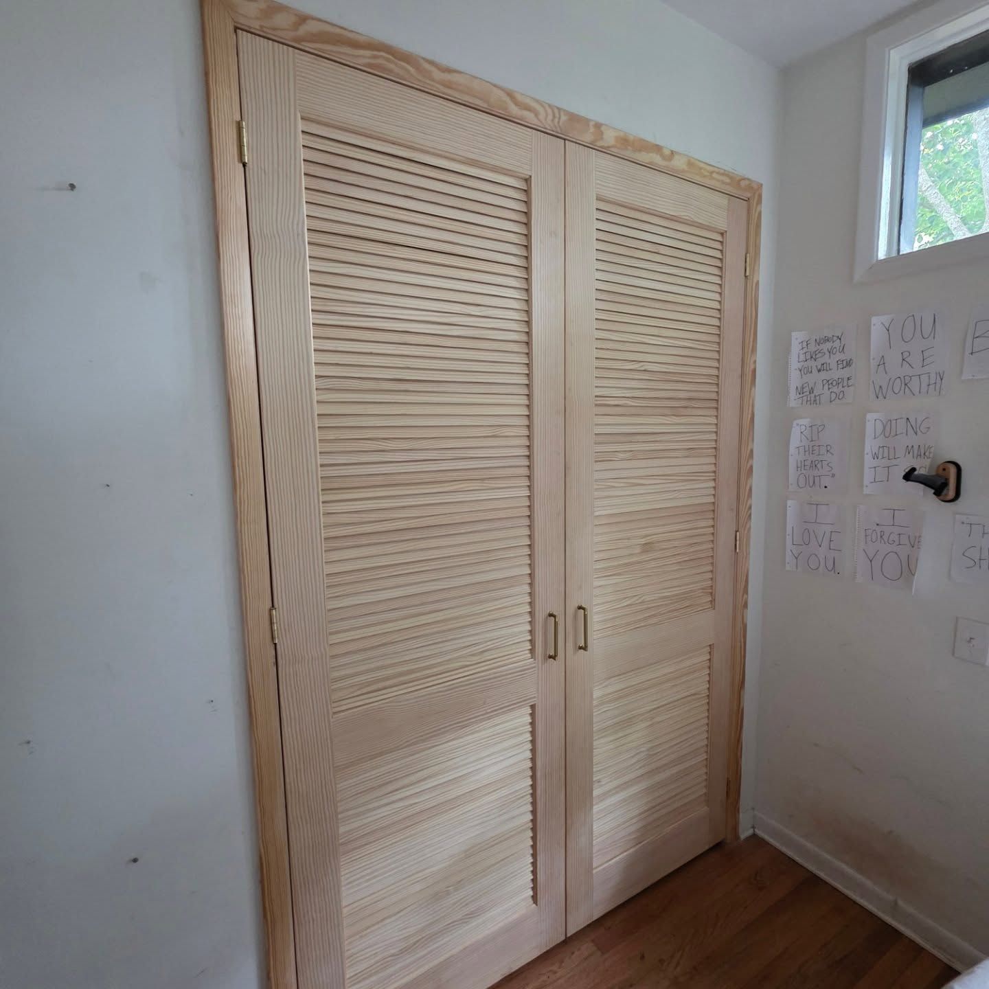 Light wood louvered closet doors in a white room with hardwood floor and wall notes.