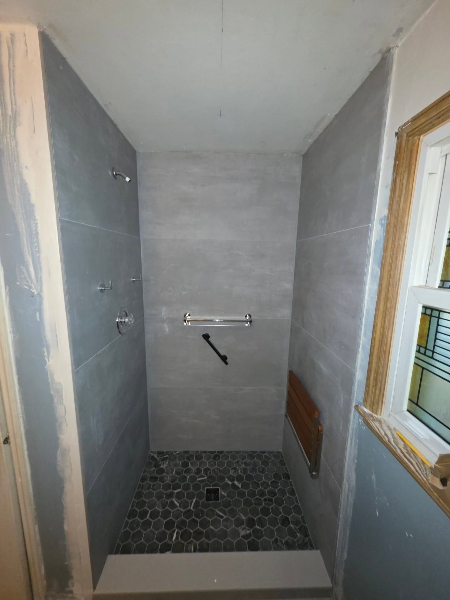 Small unfinished shower stall with gray walls, hex tile floor, and a window on the right.
