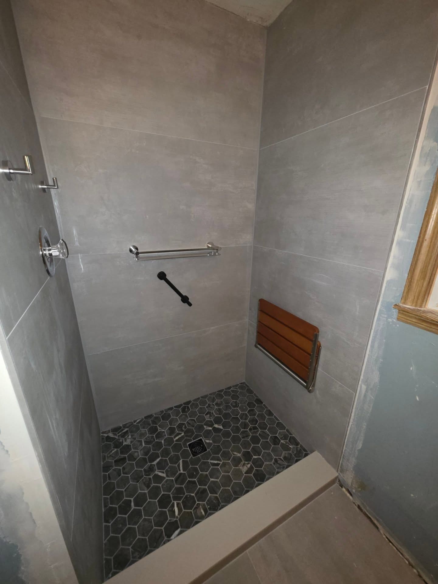 Small tiled shower stall with gray walls, hexagon floor, grab bar, and wooden bench seat