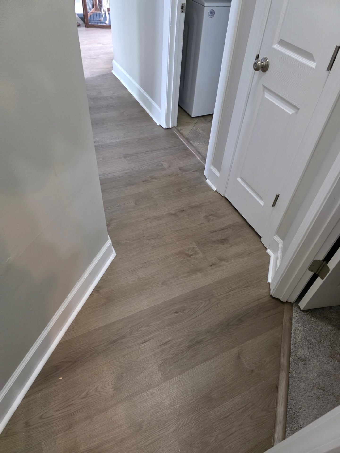 Narrow hallway with light wood flooring, white doors, and a gray carpeted edge at the right.