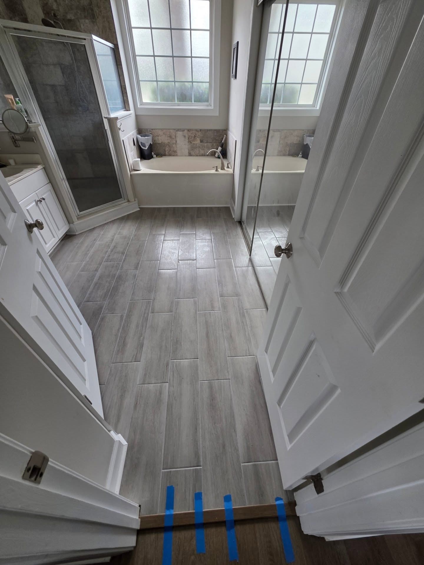 Narrow bathroom with white doors, gray tile floor, glass shower, and a bathtub by a window.