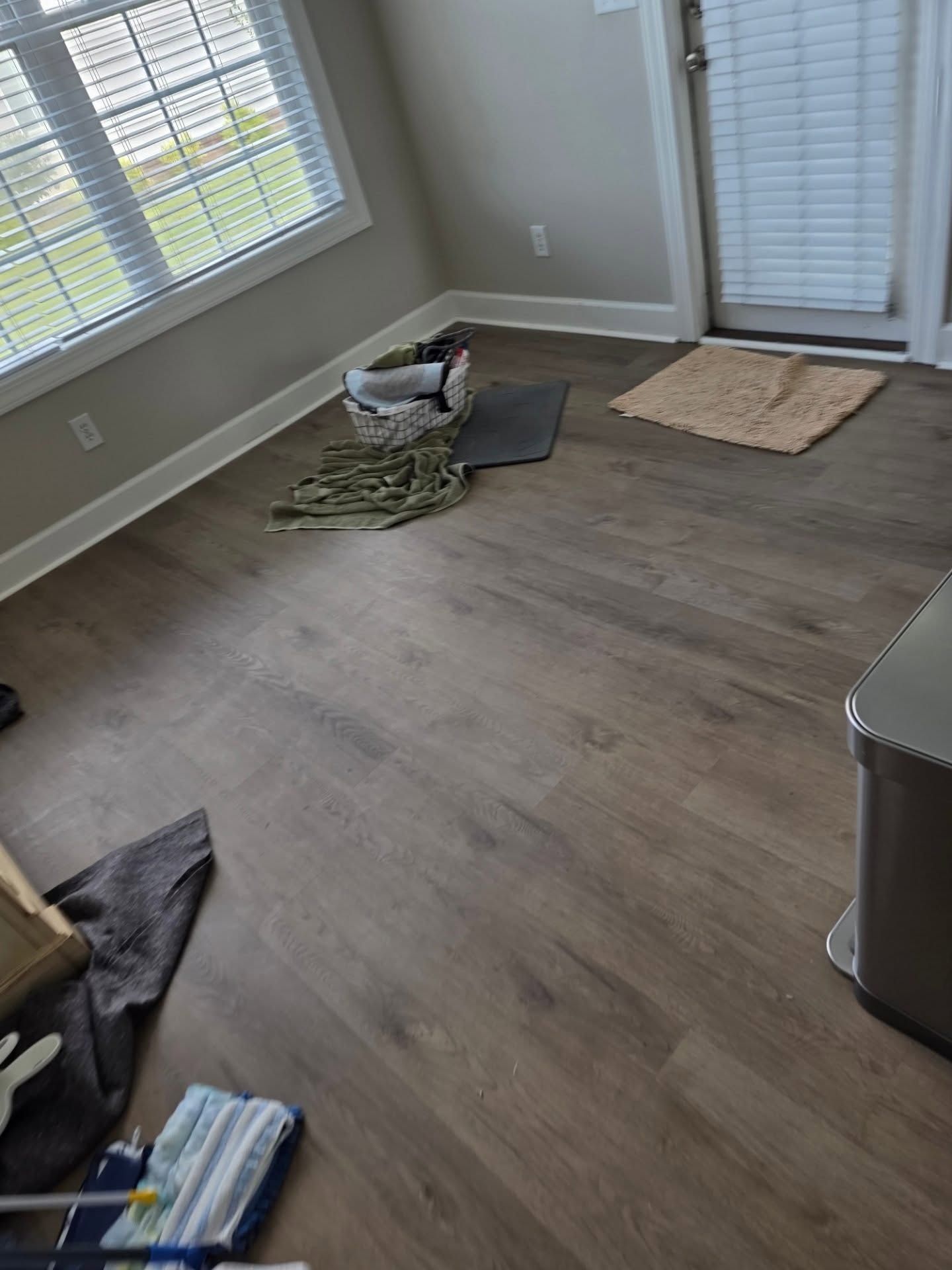 Empty living room with scattered cleaning items, gray wood floor, windows, and a beige rug by the wall.