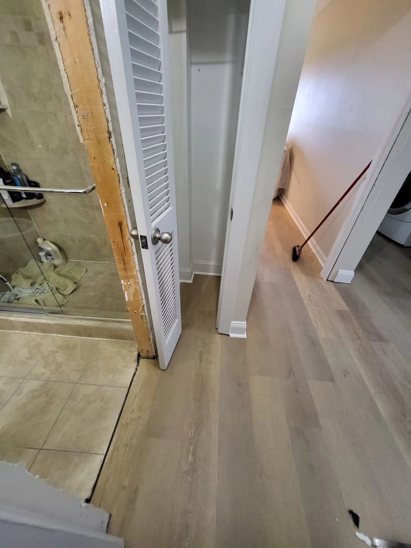 Bathroom and hallway with light wood flooring, shower on left, and louvered closet doors in center.