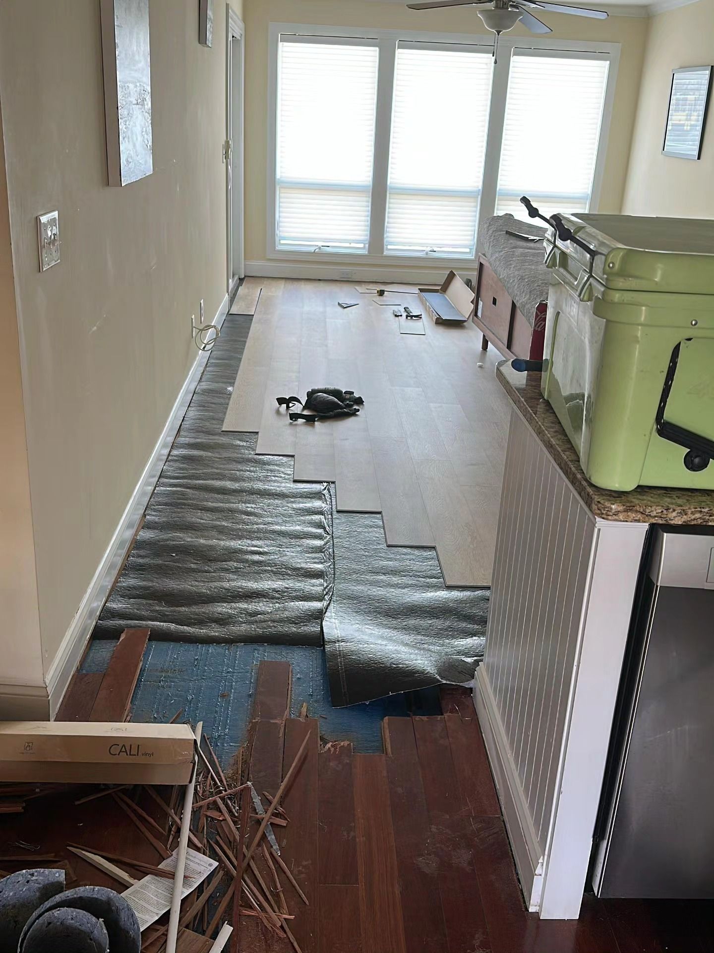 Narrow hallway with torn flooring, exposed underlayment, and construction materials near a bright windowed door