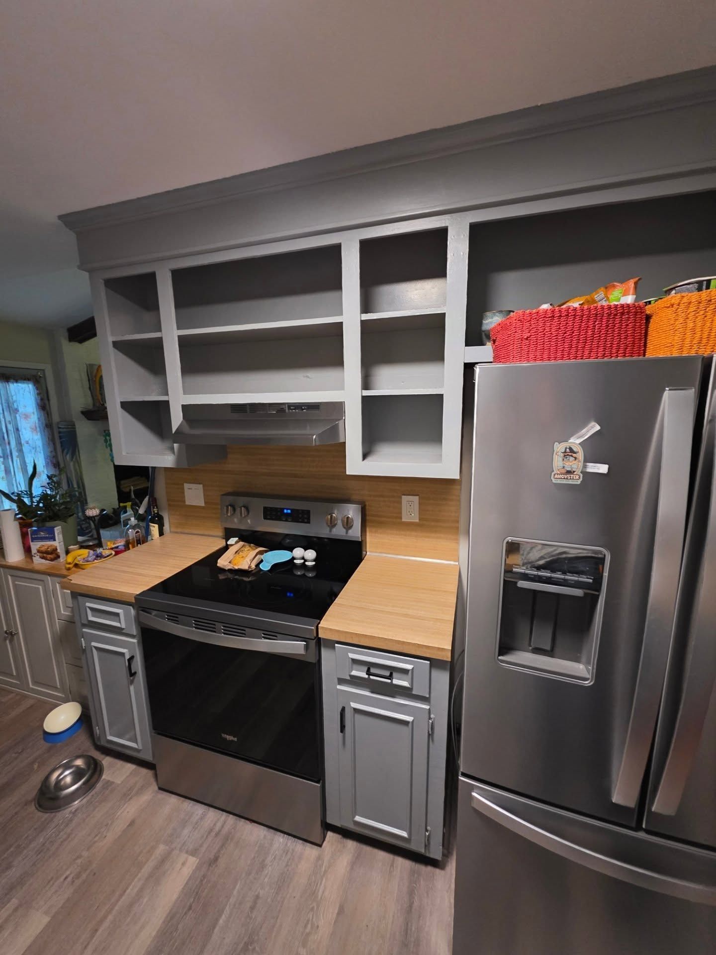Small kitchen with open gray cabinets, wood counters, black stove, and stainless steel refrigerator.