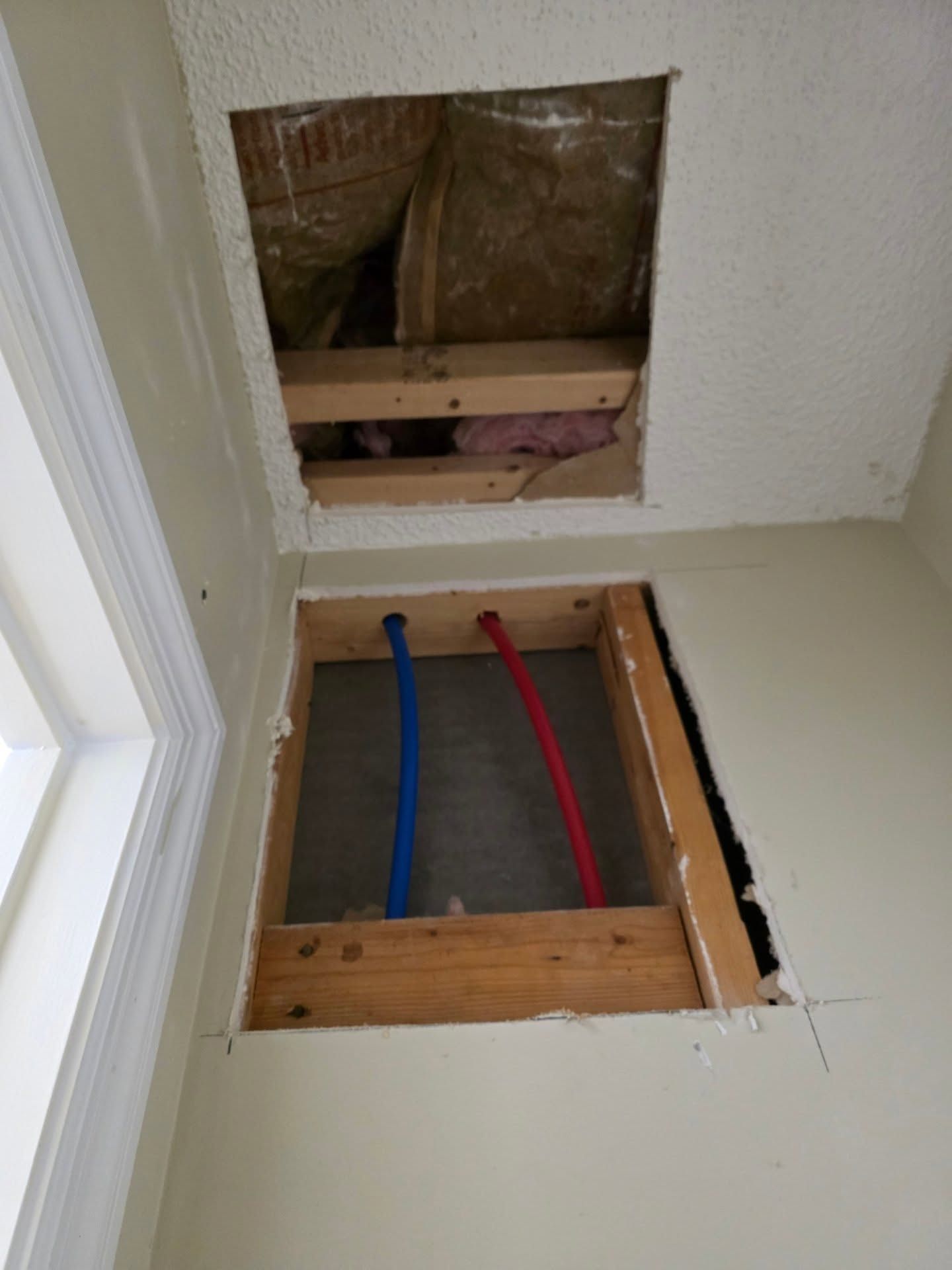 Open wall and floor access panels with exposed blue and red pipes in a room corner