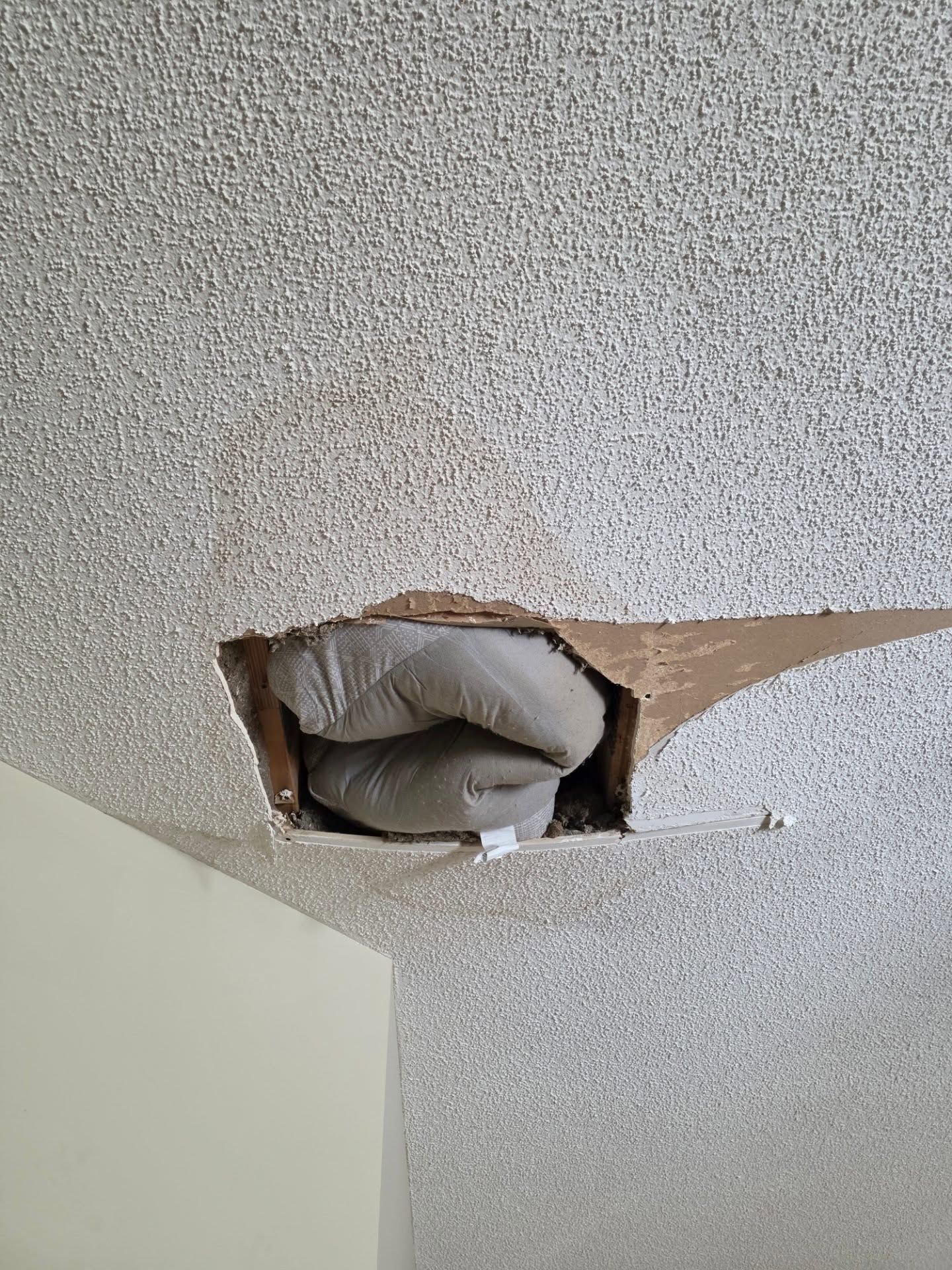 Damaged ceiling with a hole exposing white ductwork in a textured room corner