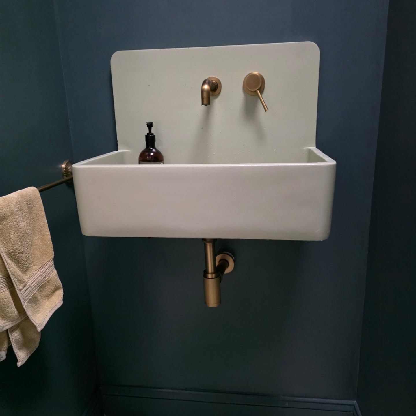 Small white wall-mounted sink with brass fixtures in a dark teal bathroom, towel hanging beside it