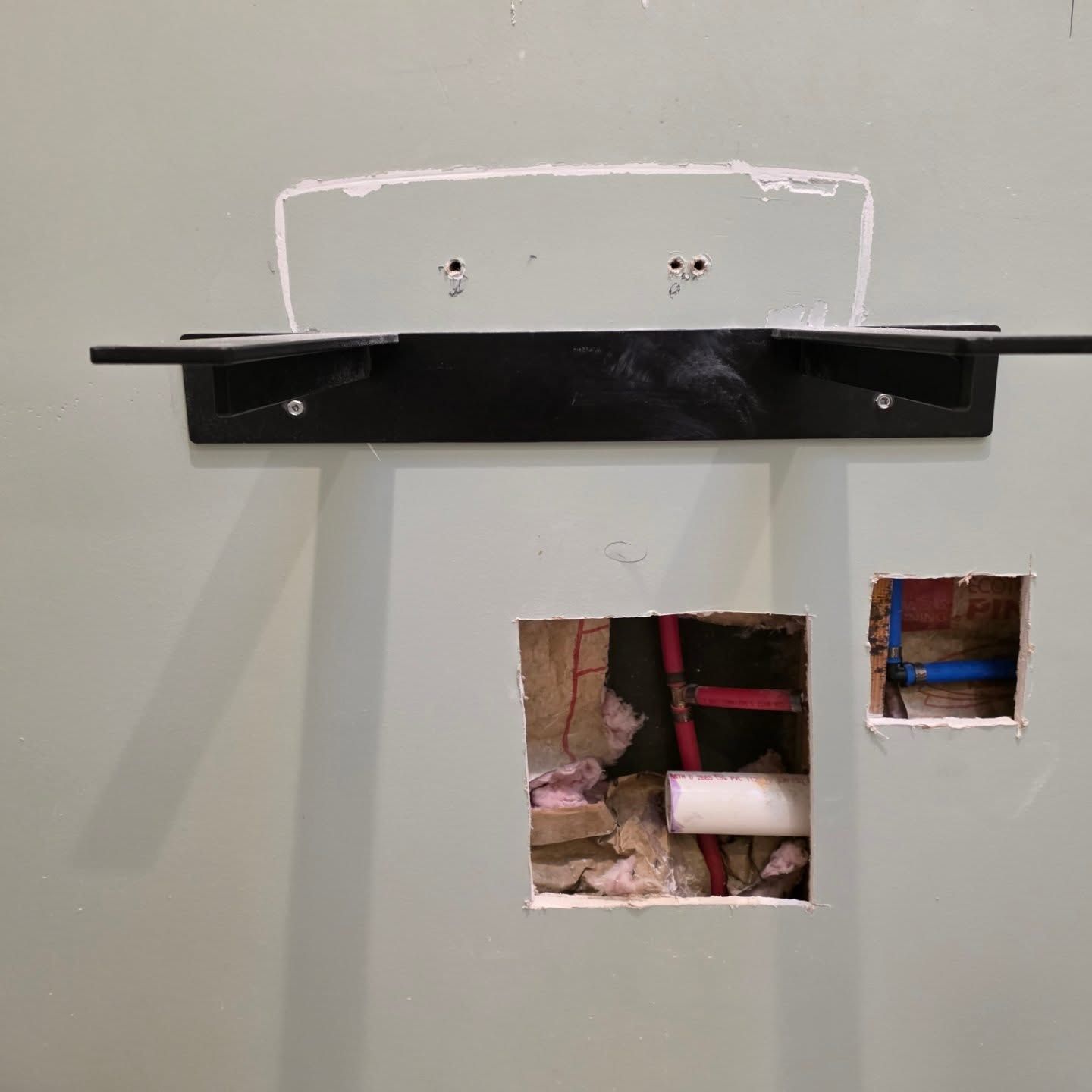 White wall with a black shelf and two cutout openings exposing plumbing behind it.