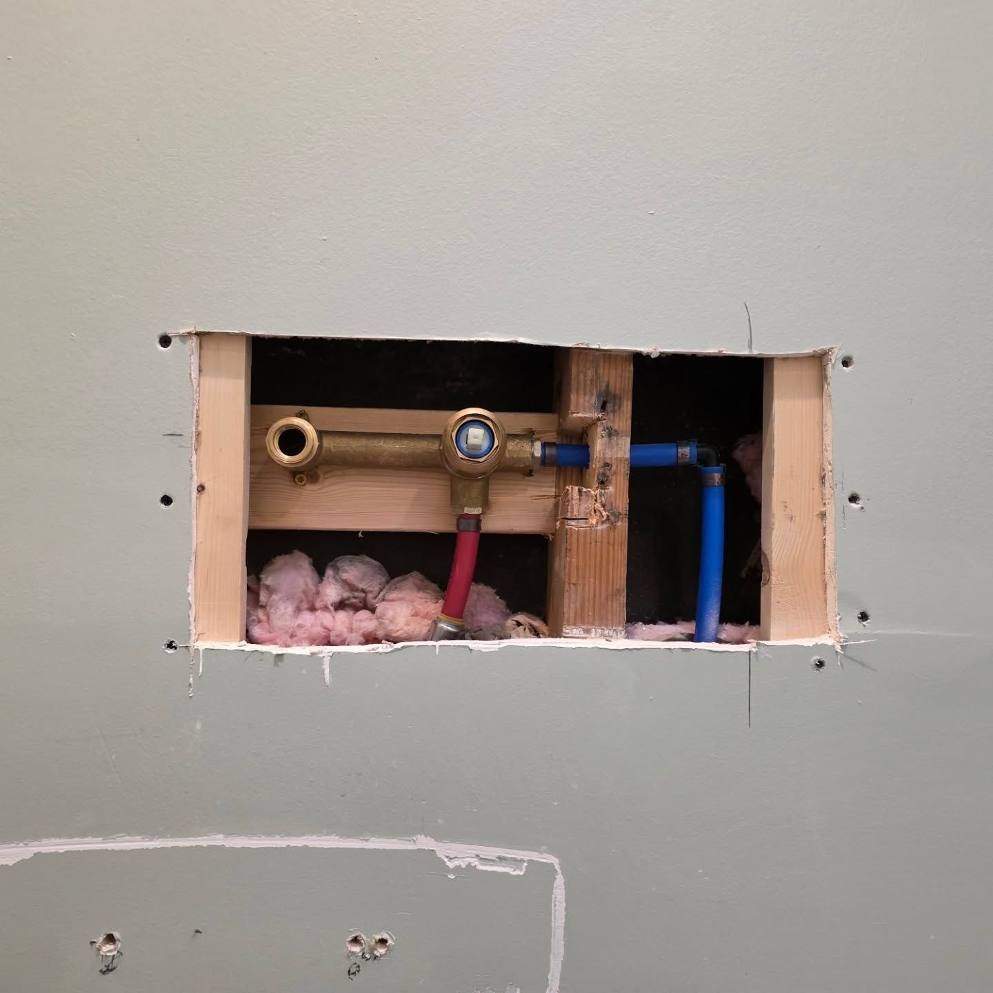 Wall cutout exposing plumbing pipes, valves, and pink insulation in an unfinished interior wall.