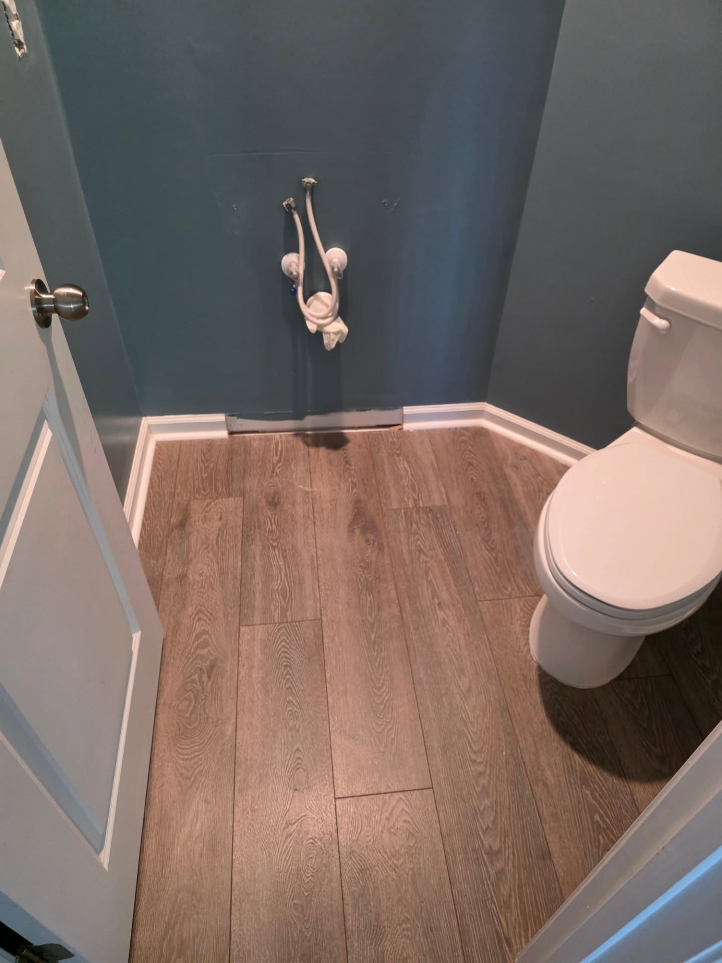 Small bathroom with wood-look floor, teal walls, toilet, and exposed plumbing at the back wall.