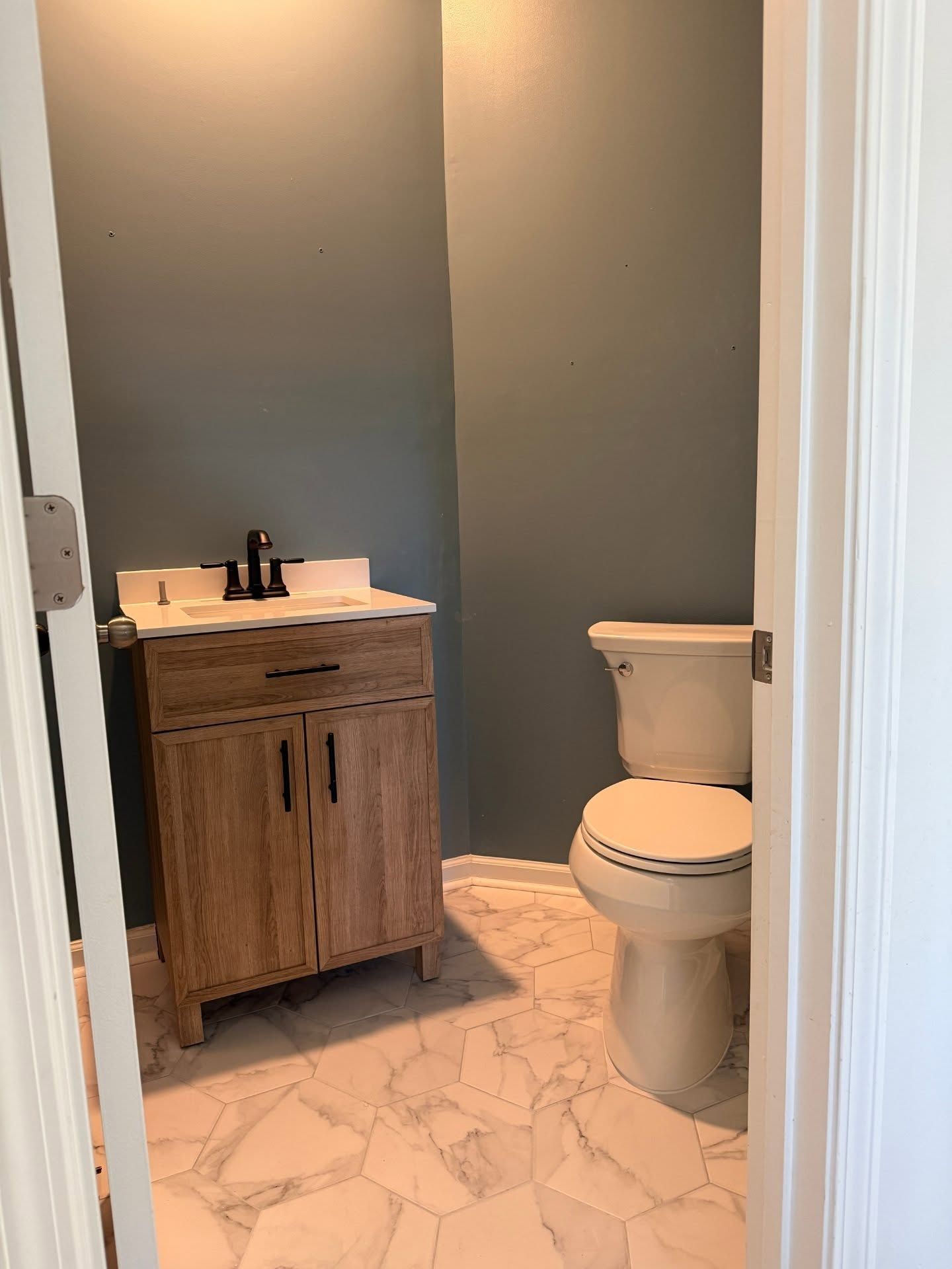 Small bathroom with gray walls, wood vanity, white toilet, and marble-pattern tile floor.