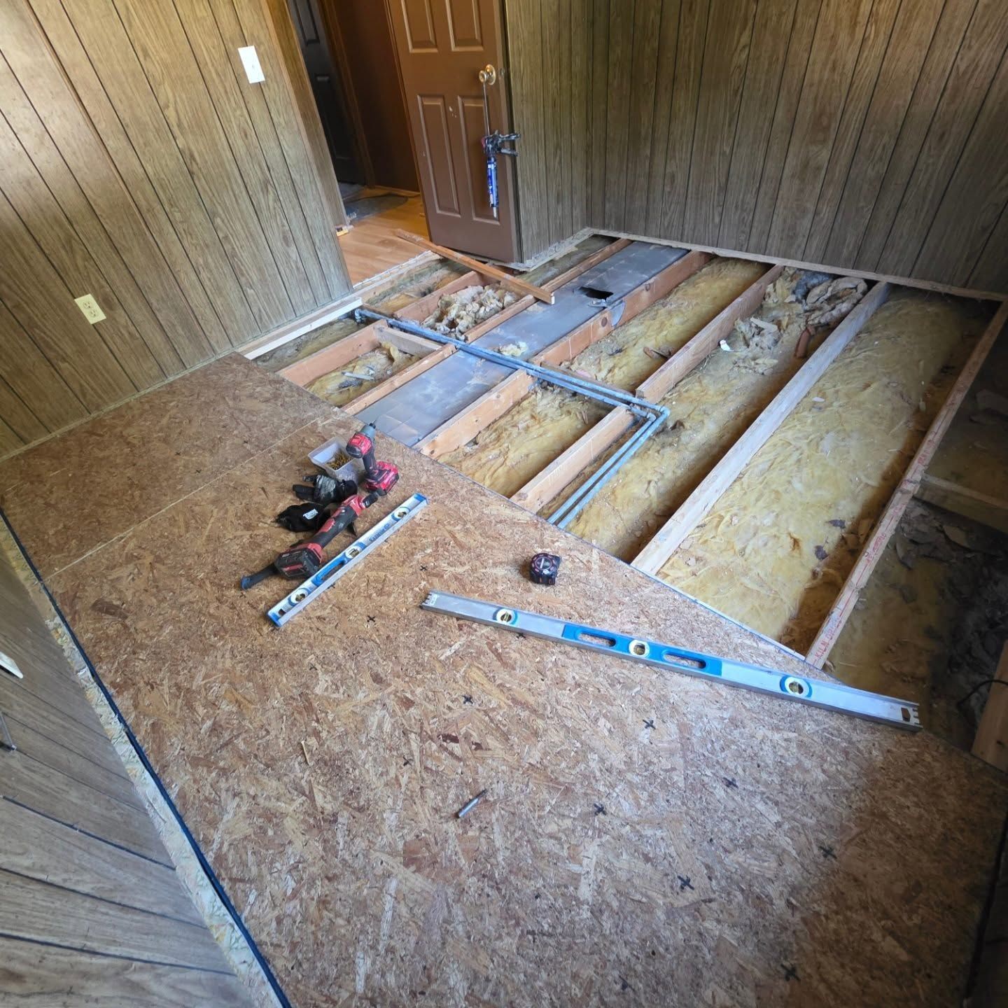 Partially demolished room with exposed floor joists, insulation, tools, and a door in the background