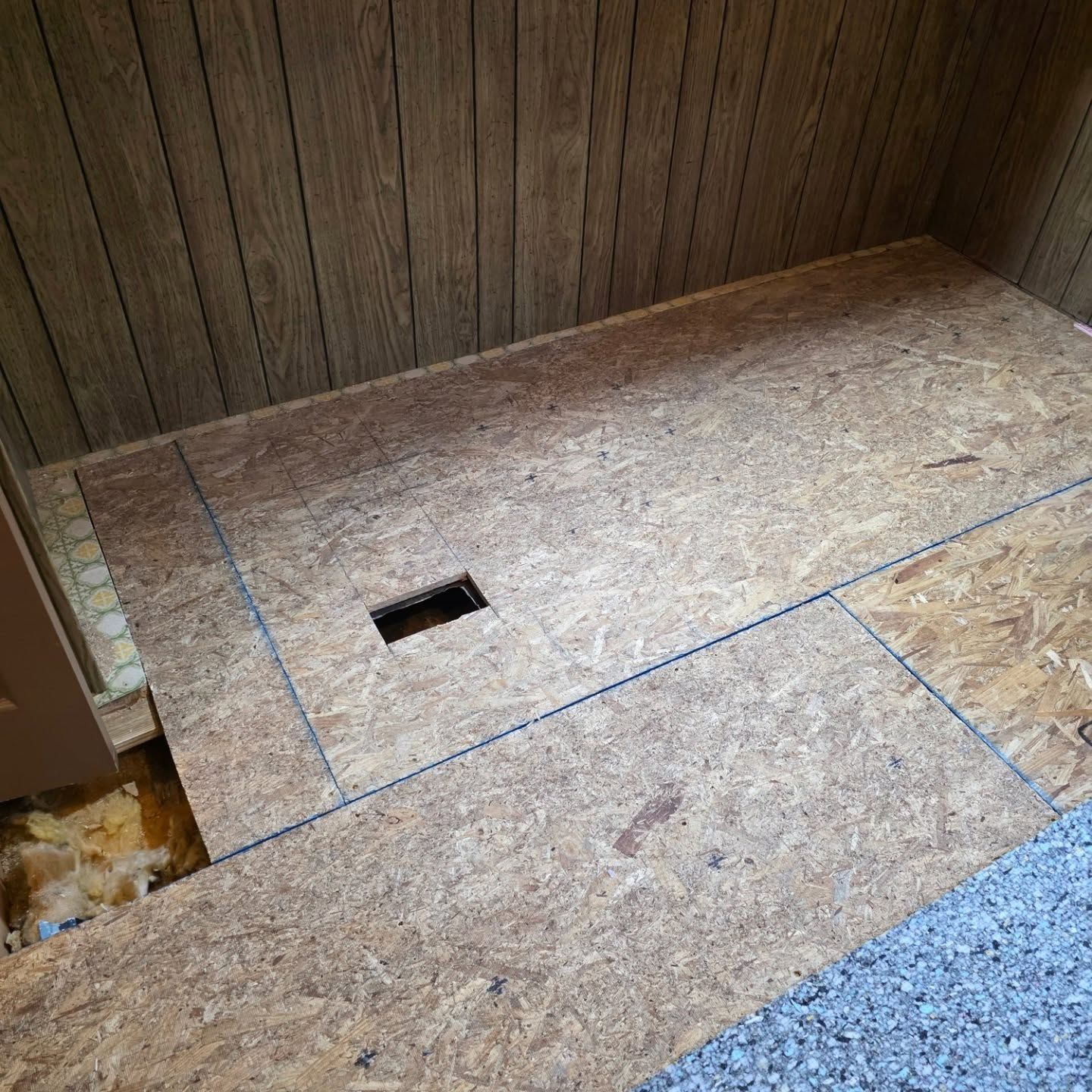 Wood-paneled room floor with exposed plywood panels and a small rectangular cutout.