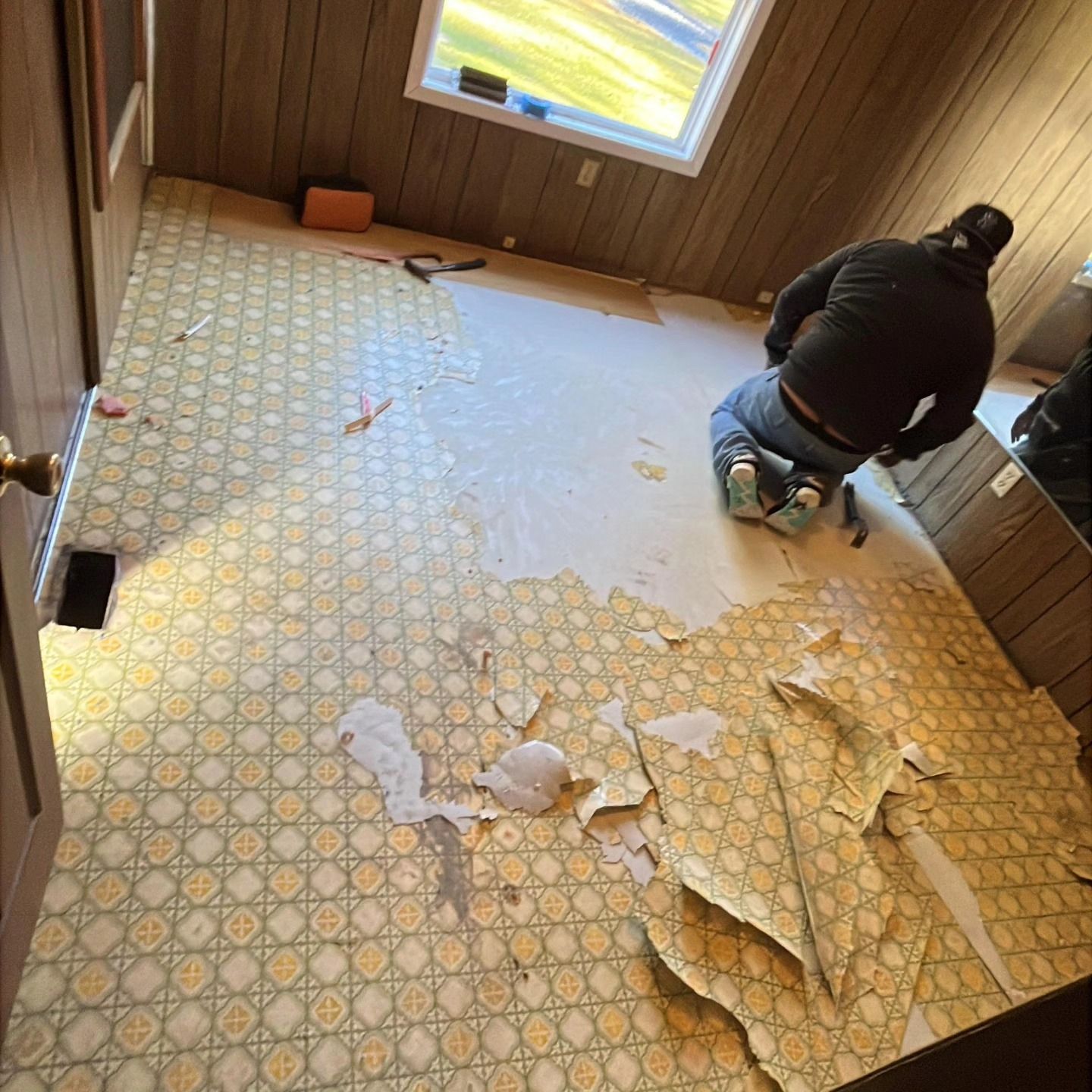 Person kneeling on torn linoleum in a room with wood-paneled walls and a window