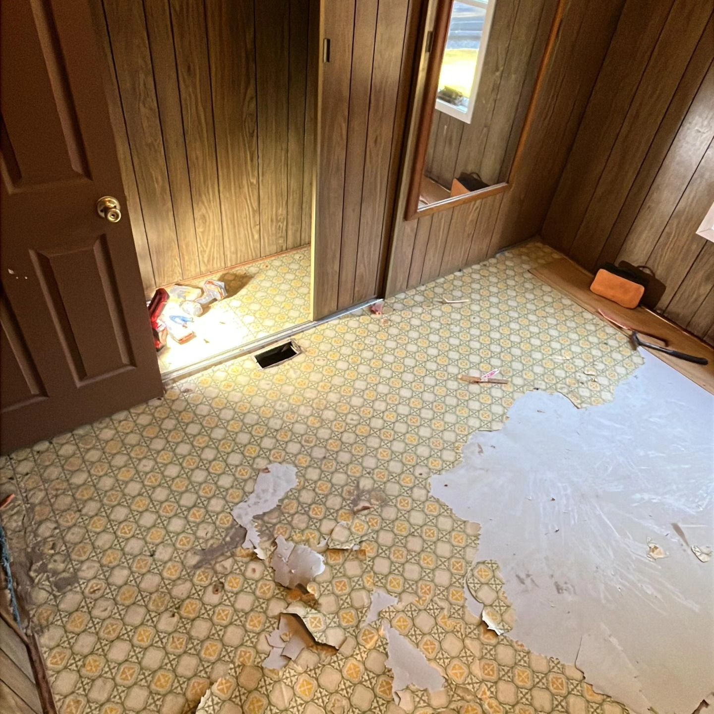 Damaged hallway floor with peeling linoleum, wooden walls, and a partially open door with daylight spilling in