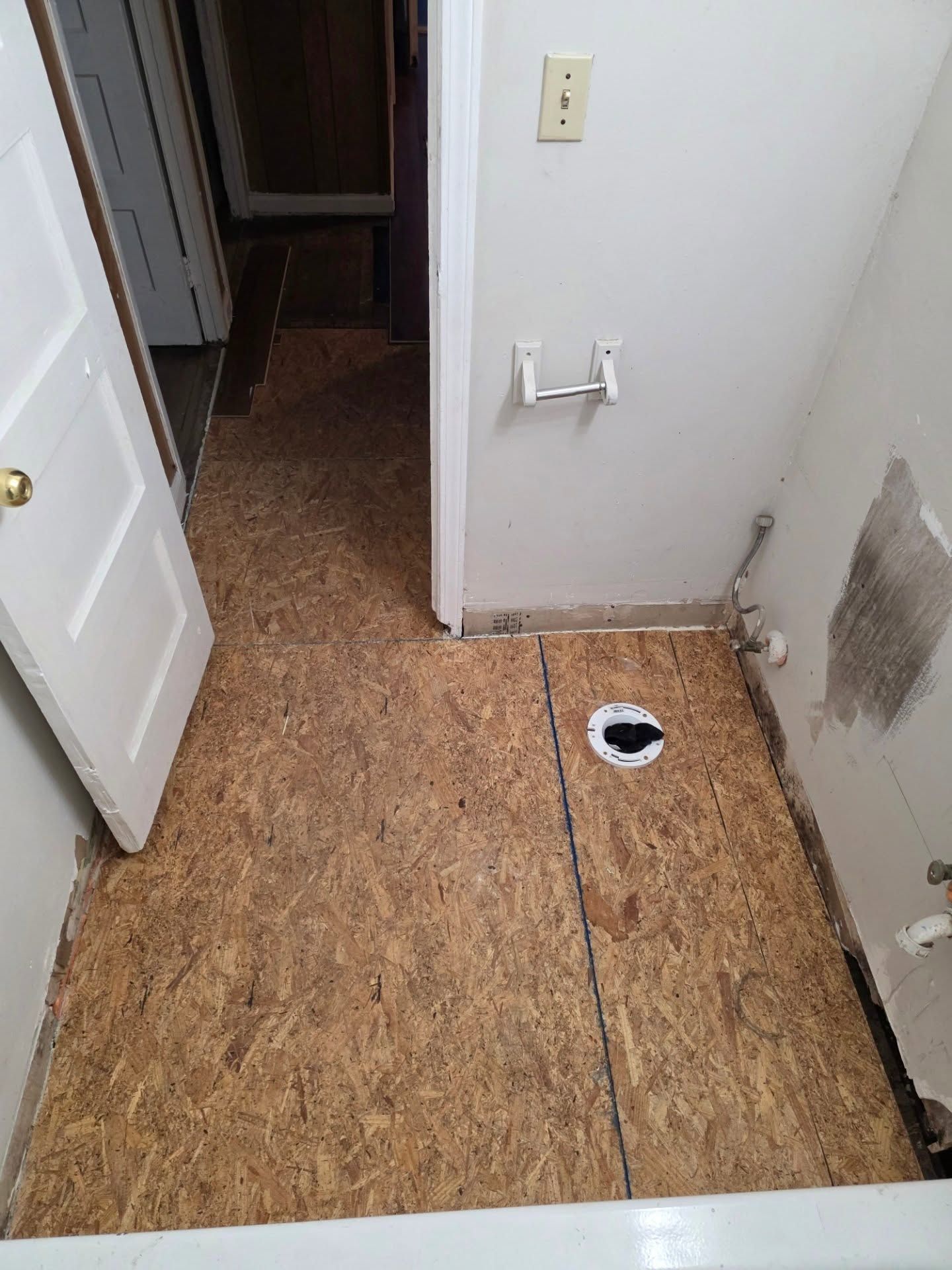Small utility room with plywood subfloor, wall plumbing outlet, and a floor drain opening.