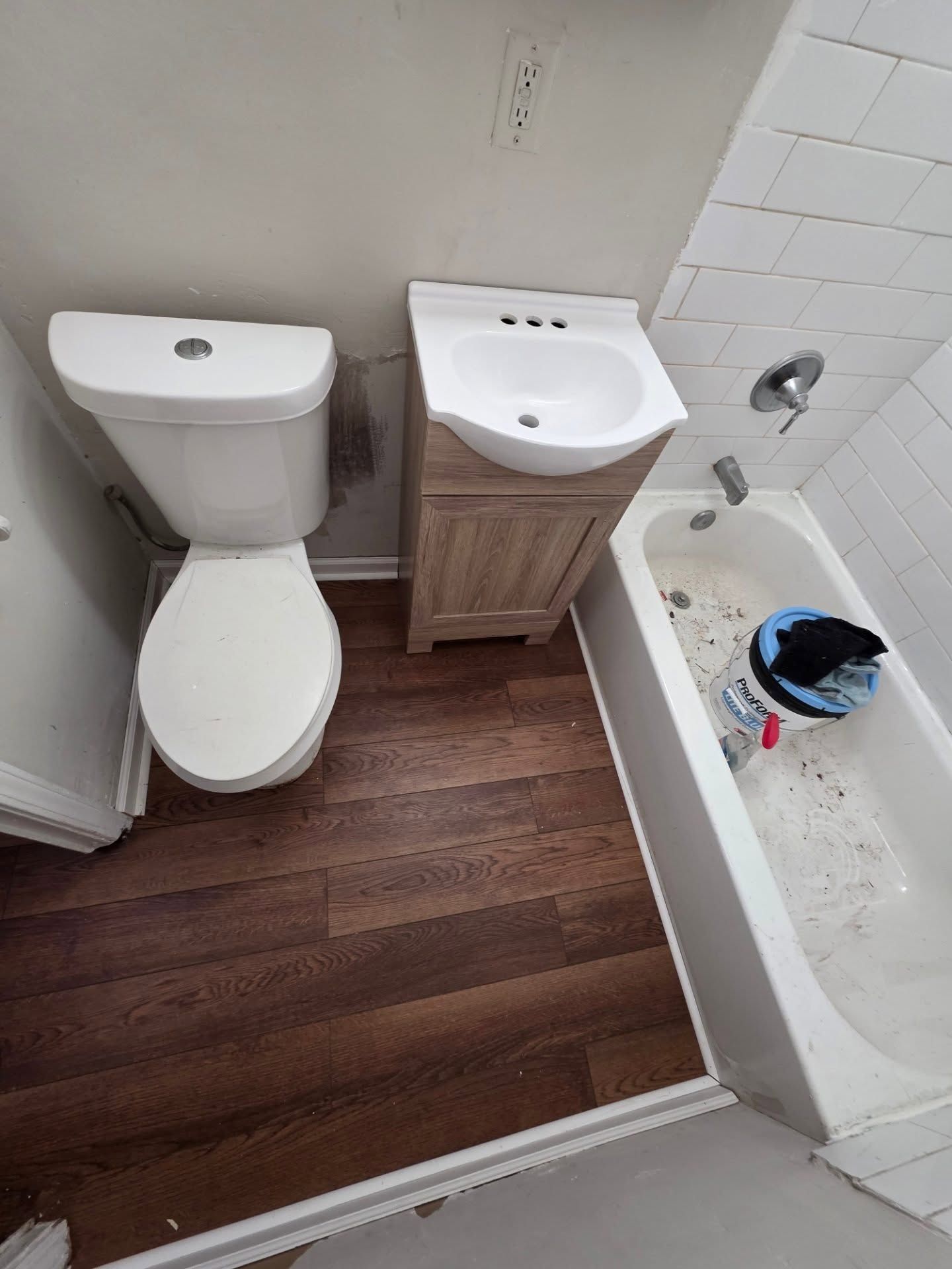 Small bathroom with white toilet, sink cabinet, bathtub, and wood-look floor.