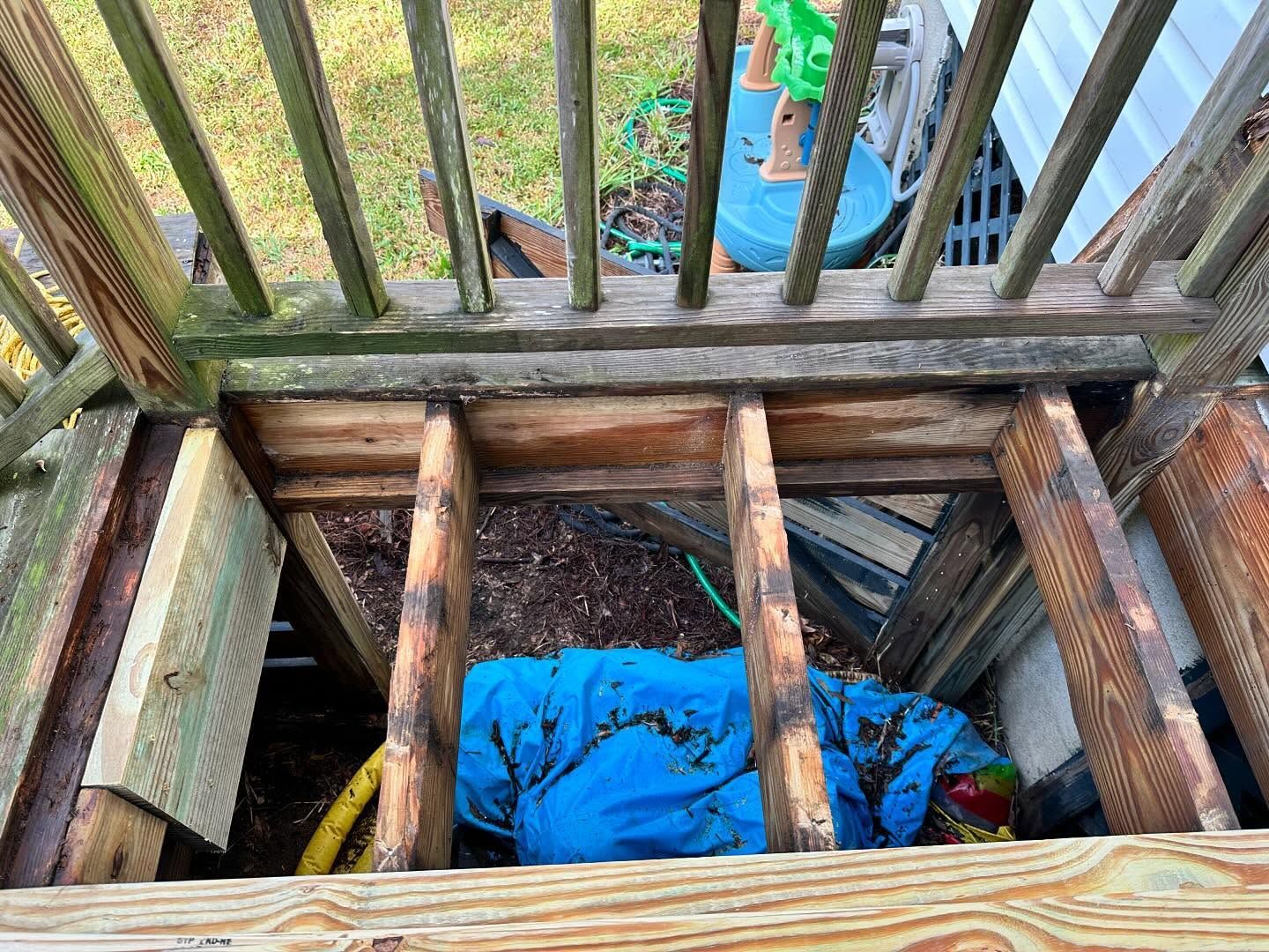 Blue tarp covering a hole beneath a wooden deck, with beams and railings overhead