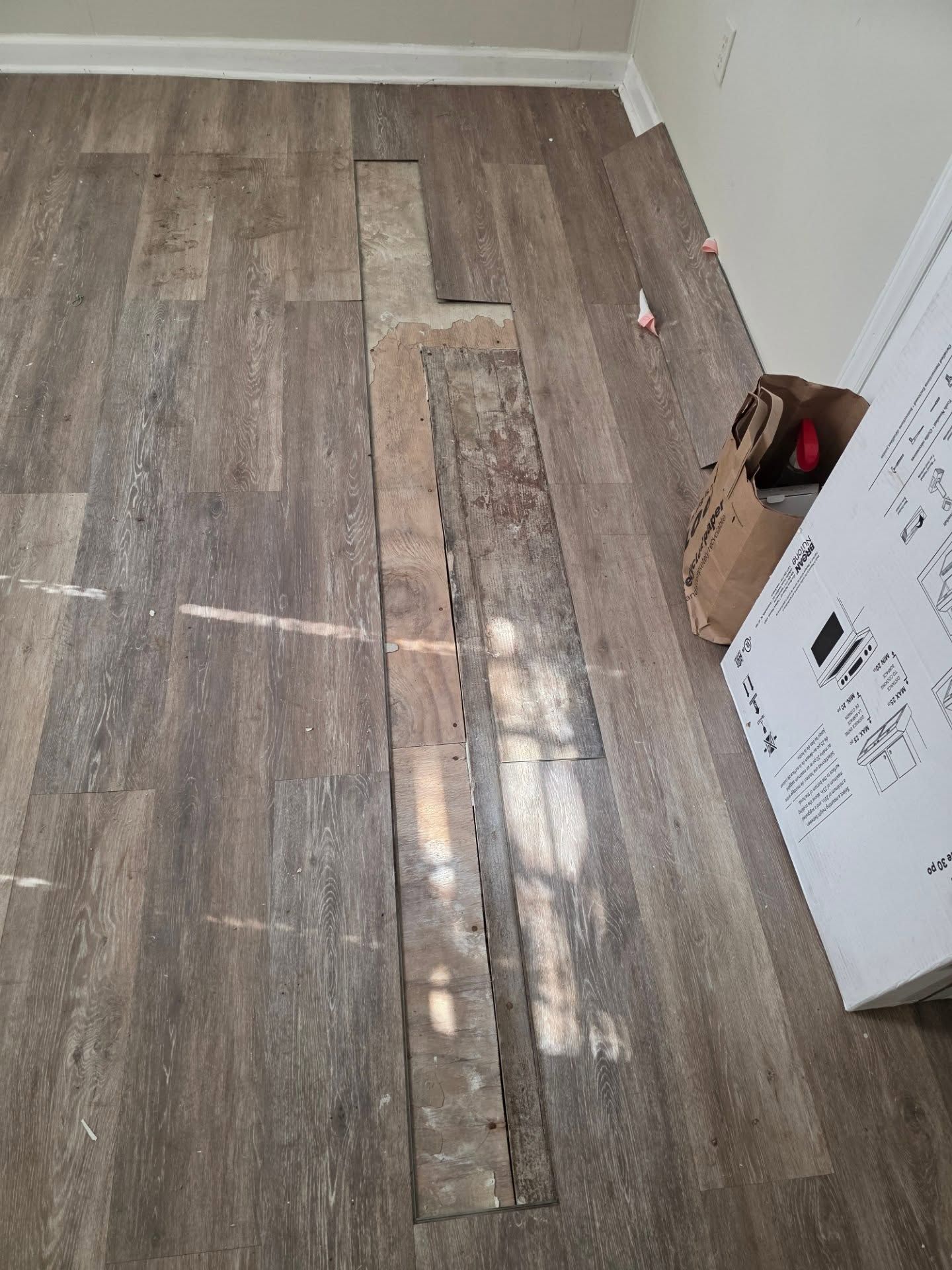 Narrow hallway with gray wood-look flooring, a damaged center plank, and boxes along the right wall.