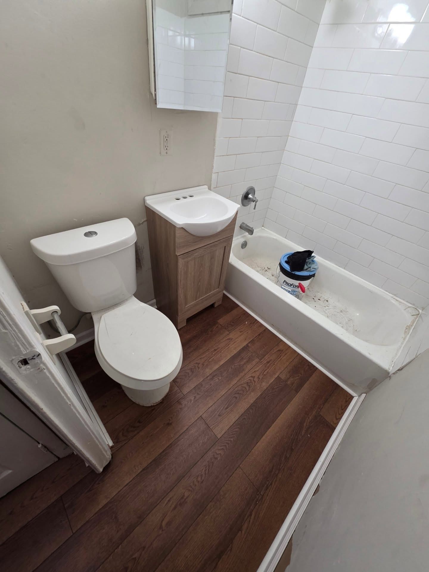 Small bathroom with toilet, sink vanity, and bathtub; dark wood floor and white tiled walls.