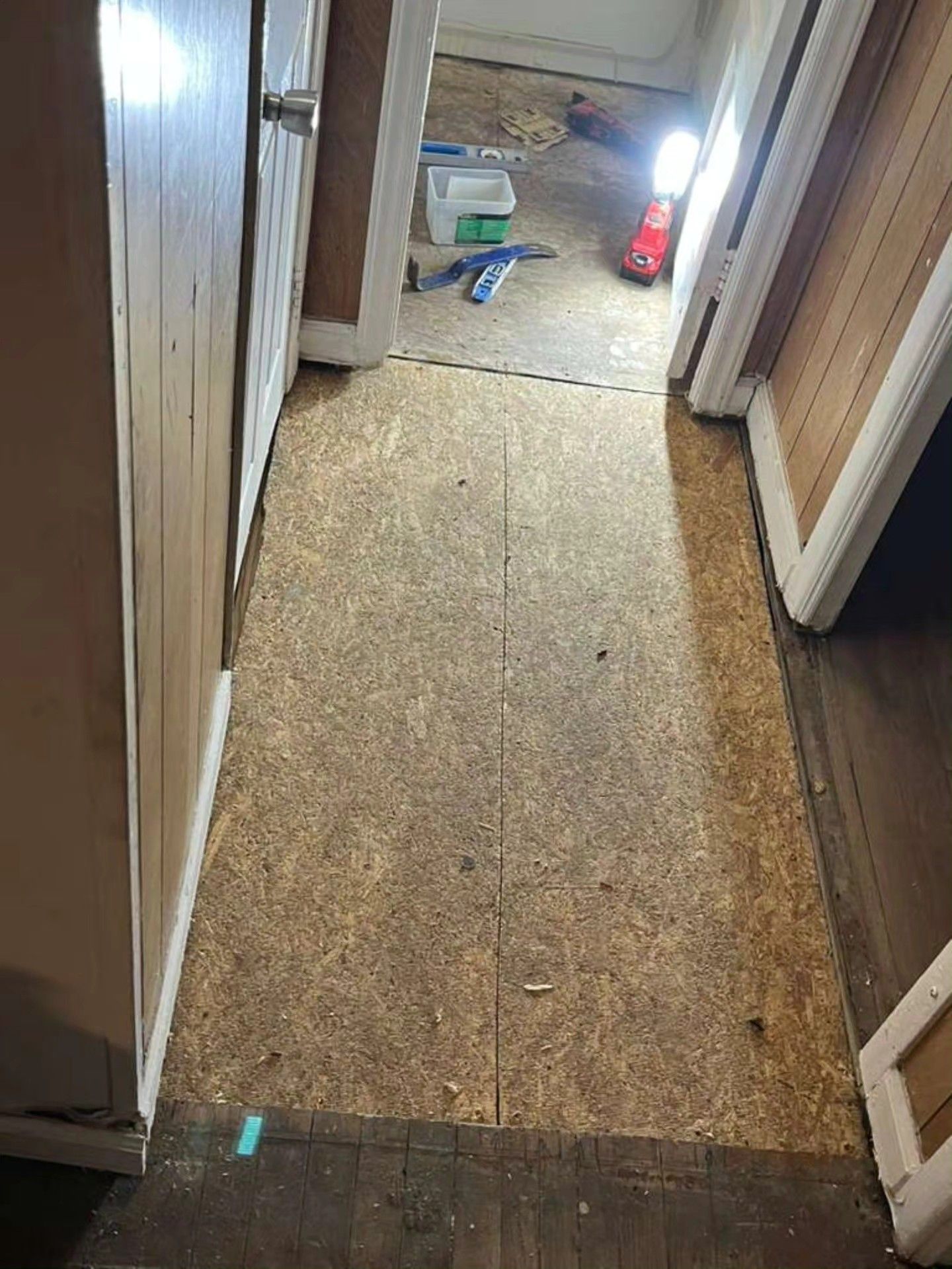 Narrow hallway with unfinished floor leading to a cluttered room with a small red light in the back.